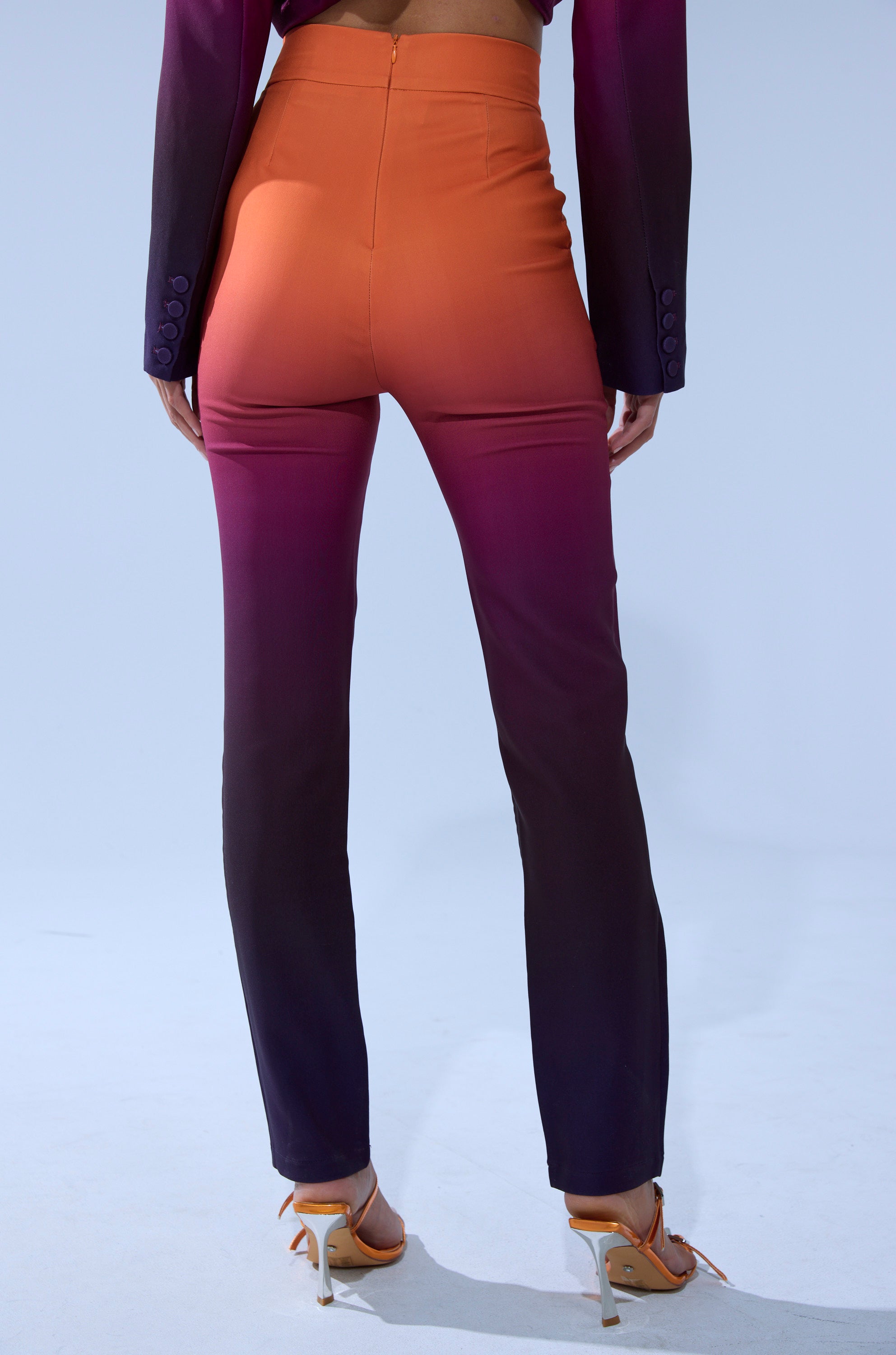 A person faces away wearing the RISE AND SHINE TROUSER, a high-rise pant with an orange-to-deep purple gradient fade, paired with orange heeled sandals. The background is plain and light-colored.