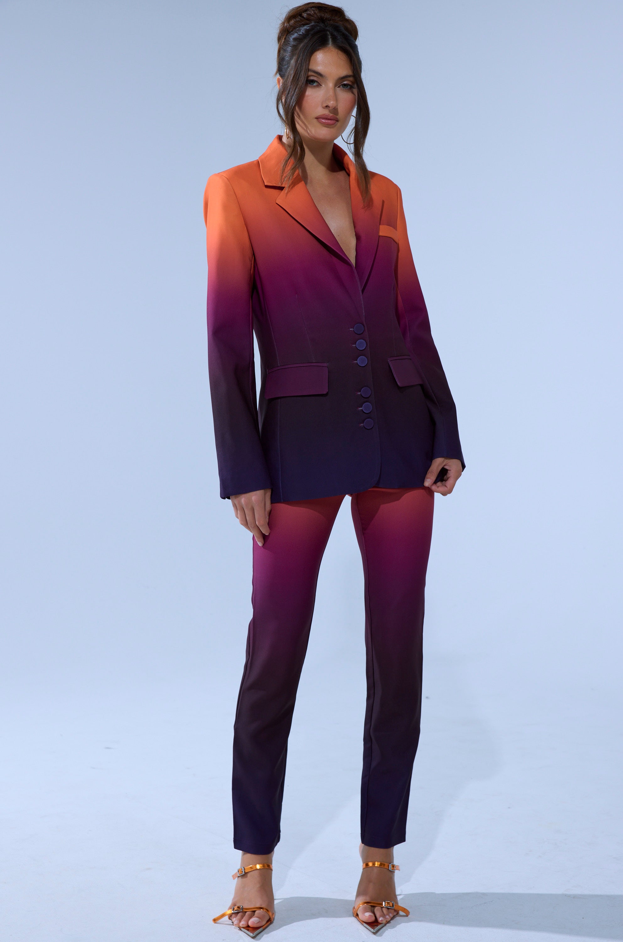 A woman stands confidently in the RISE AND SHINE TROUSER and an ombre blazer that fades from orange at the shoulders to deep purple. Her long hair is styled up, she wears gold open-toe heels, and the background is plain and light-colored.