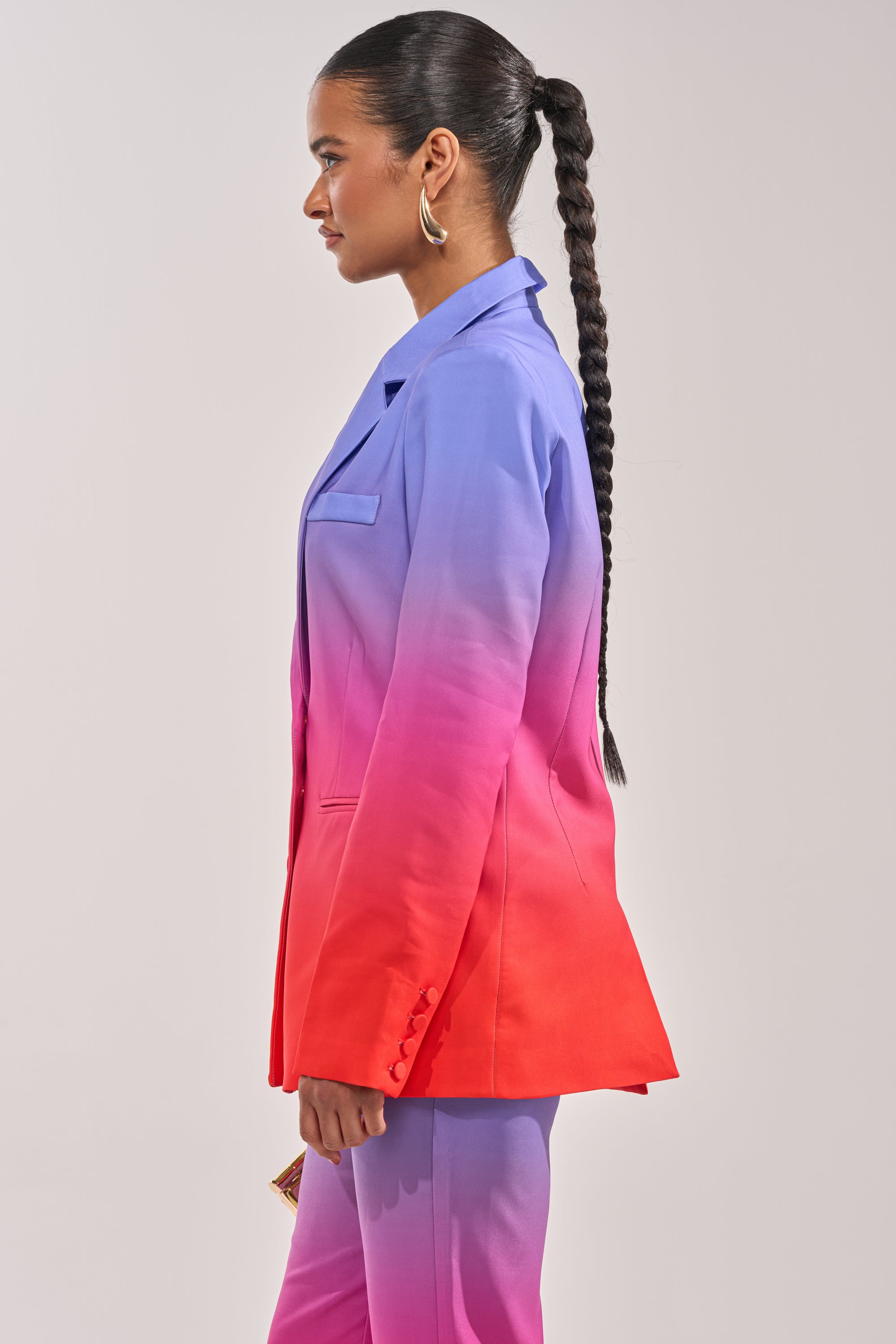 A woman in profile wears the SUNSET IN PARADISE OMBRE BLAZER, showcasing a vivid blue-to-pink gradient. She sports a long braided ponytail and gold hoop earrings, set against a simple light background. 3