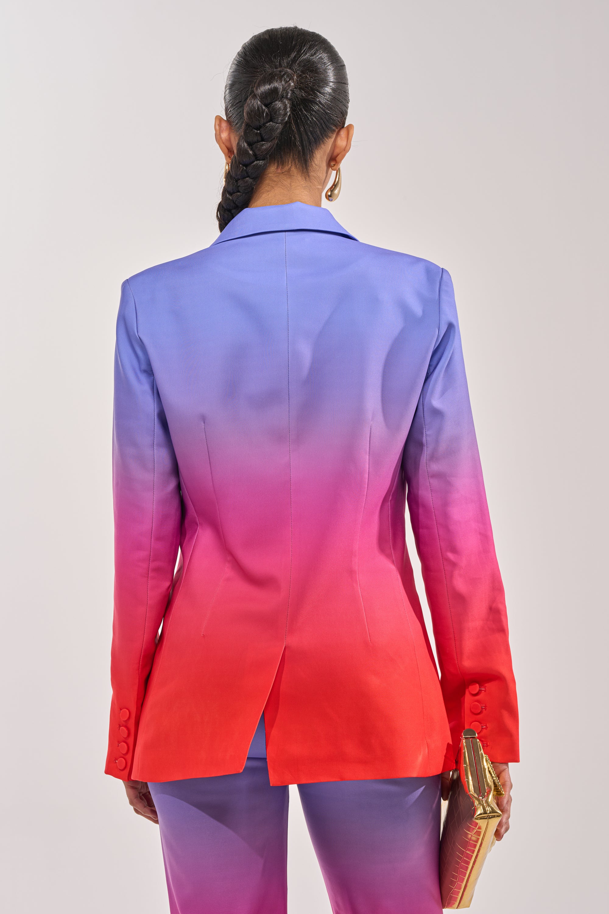 A person with a long braid stands facing away, wearing the SUNSET IN PARADISE OMBRE BLAZER—blue at the shoulders fading to pink and bright red at the hem—and holds a gold clutch in their left hand. 4