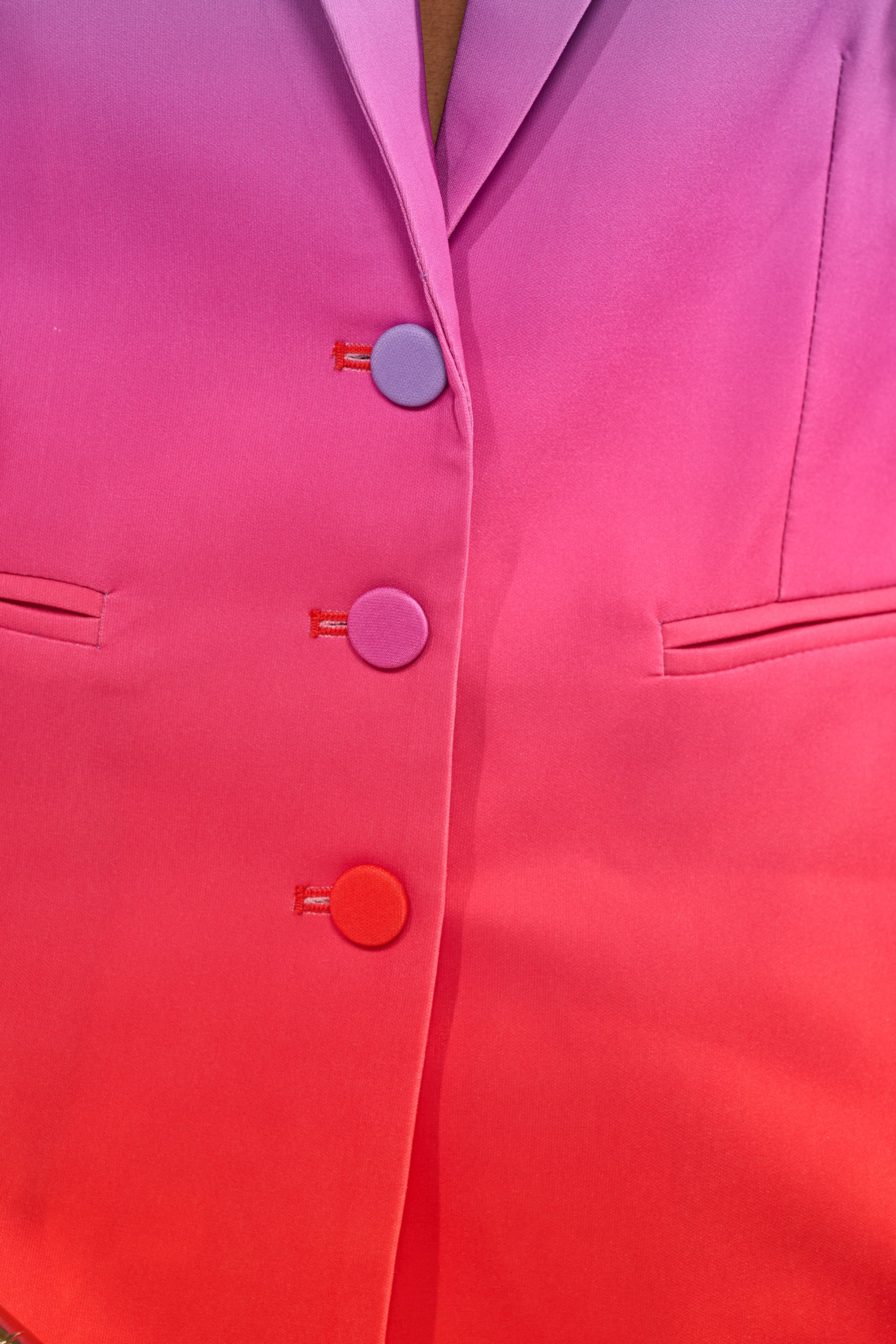 Close-up of the SUNSET IN PARADISE OMBRE BLAZER, a tailored women's jacket with a pink-to-red ombré gradient, three matching round buttons, and two horizontal slit pockets. 7