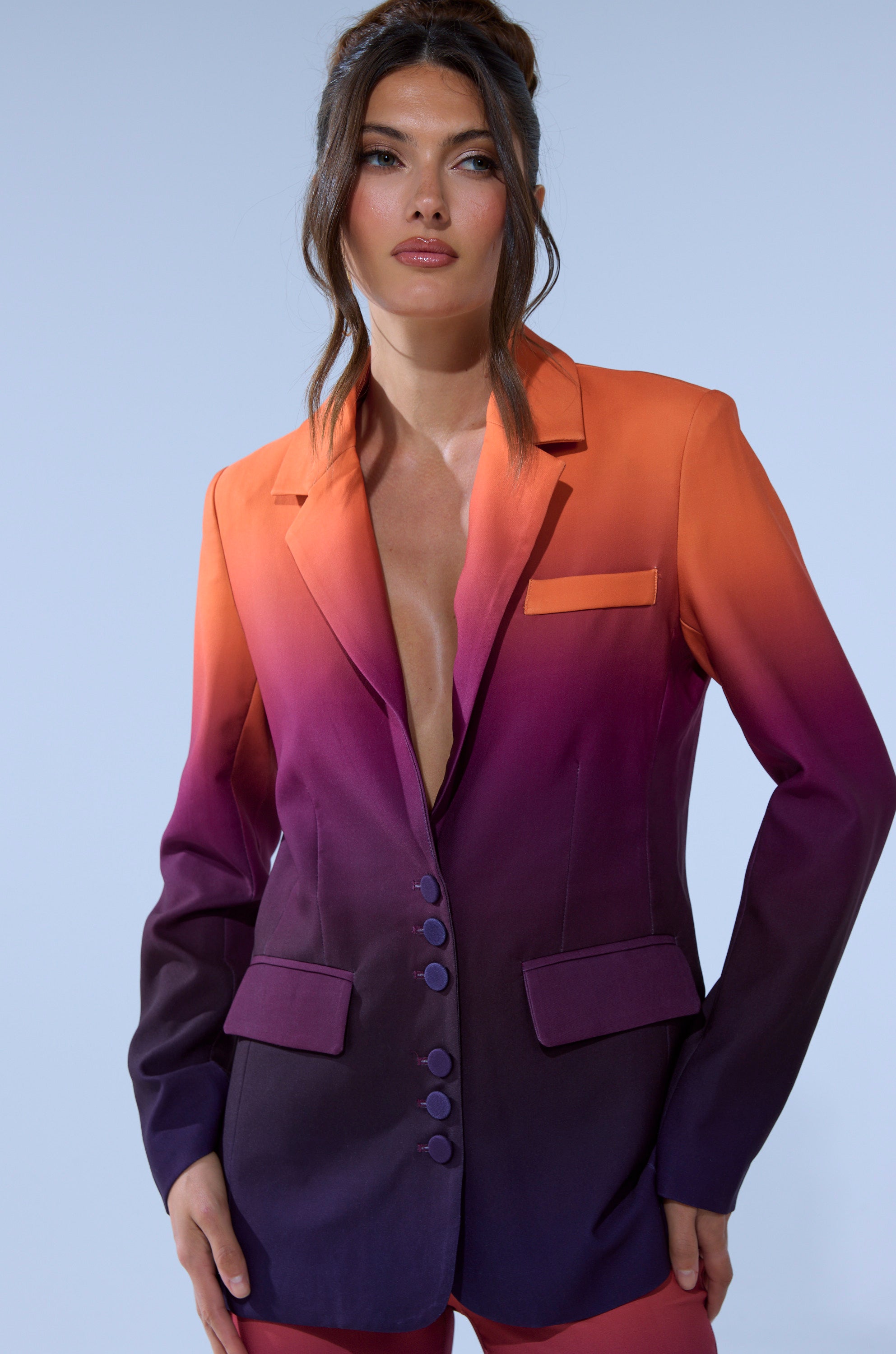 A woman poses against a light blue backdrop wearing the RISE AND SHINE OMBRE BLAZER, a tailored jacket with buttons and pockets. The blazer features an orange-to-deep-purple ombre, creating a striking sunset effect from shoulders to hem.