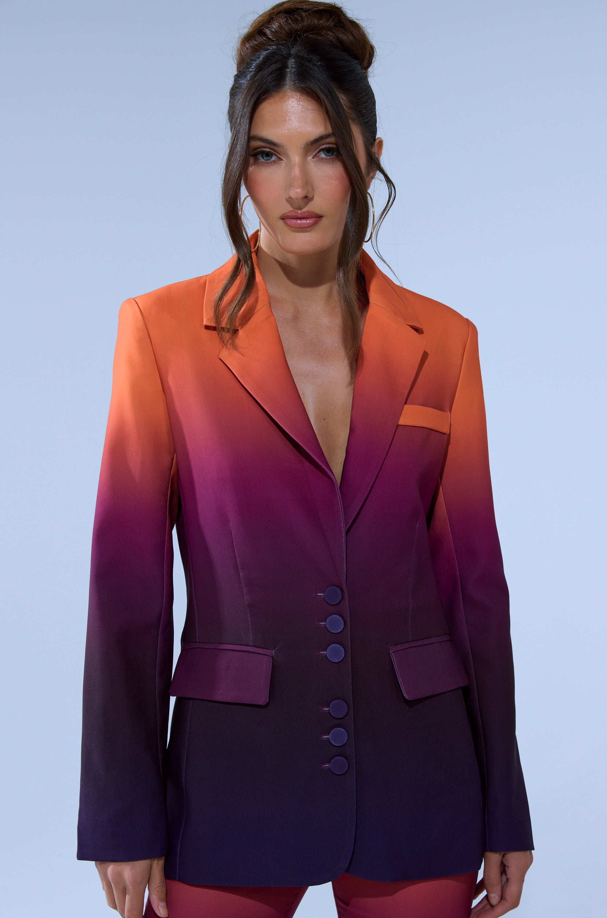 A woman with long brown hair in an updo wears the RISE AND SHINE OMBRE BLAZER, a tailored blazer with an orange-to-deep purple fade, over matching pants, standing against a plain light background.