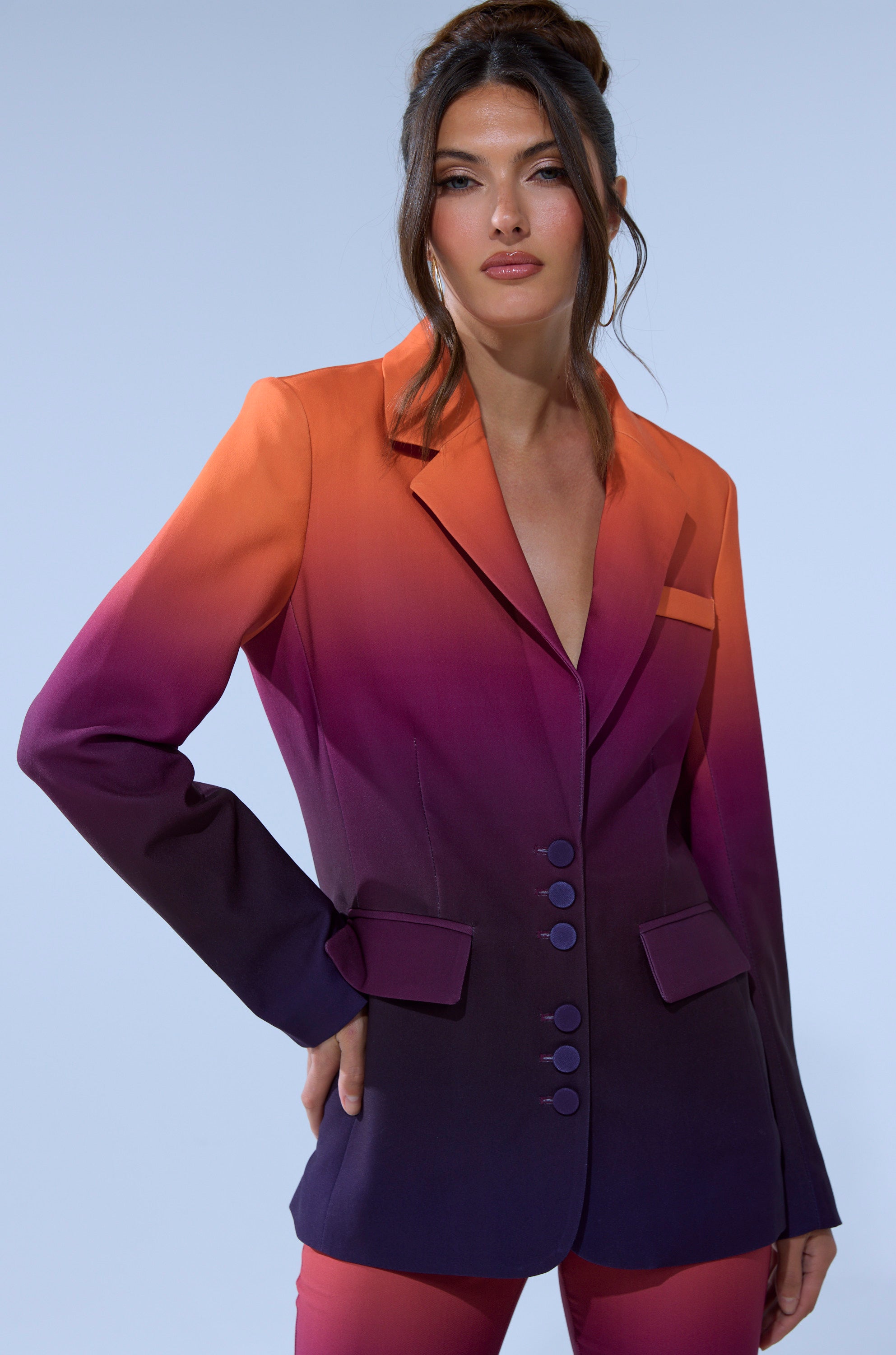 A woman wears the RISE AND SHINE OMBRE BLAZER, its sunset-inspired gradient shifting from orange to deep purple, paired with matching pants against a light blue backdrop.