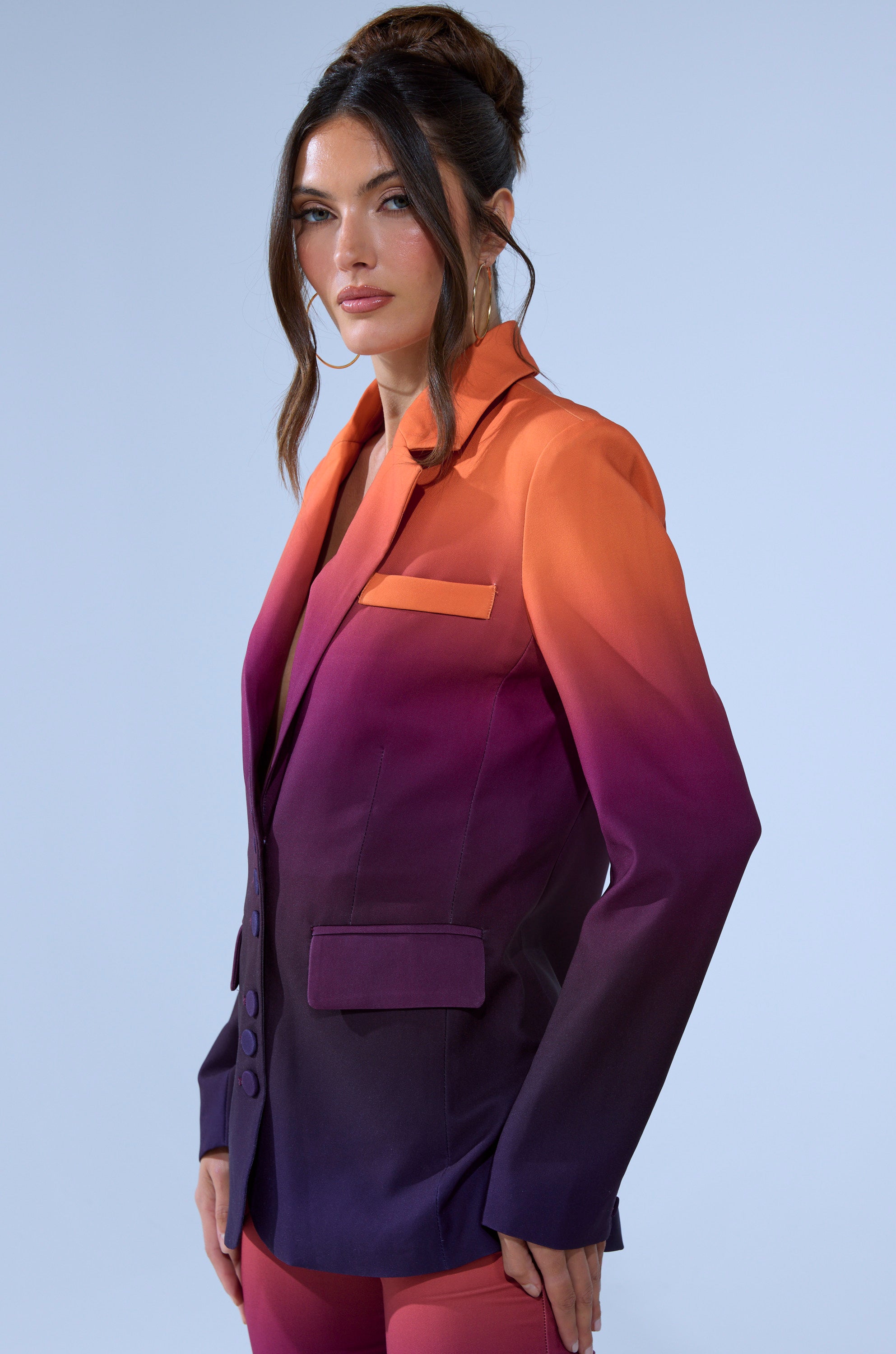 A woman with long brown hair in an updo wears the RISE AND SHINE OMBRE BLAZER, which features an ombre color gradient from orange to deep purple, set against a plain light blue background.