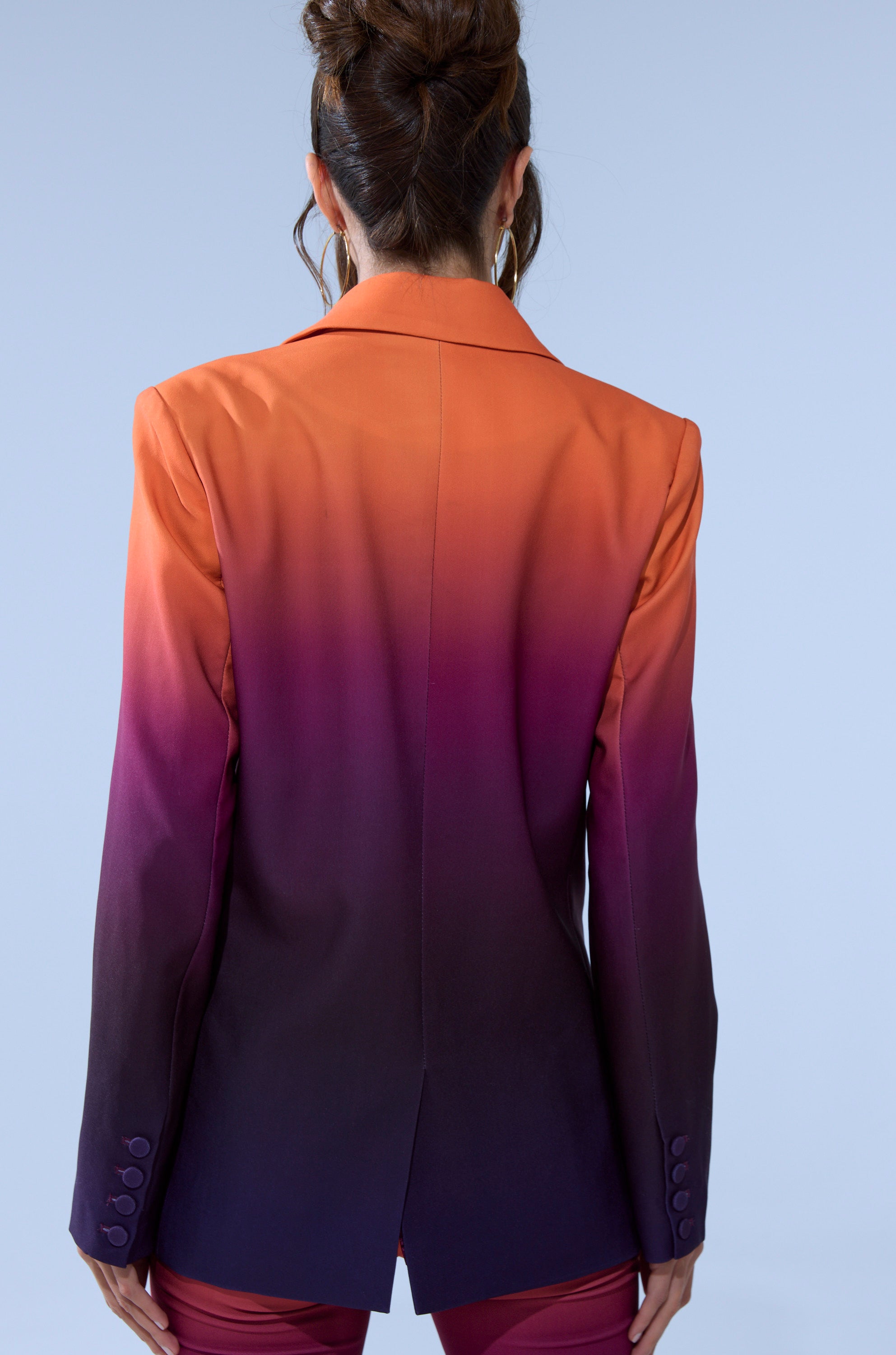 A person with dark hair in an updo is shown from behind, wearing the RISE AND SHINE OMBRE BLAZER with an orange-to-purple gradient, set against a light blue background.