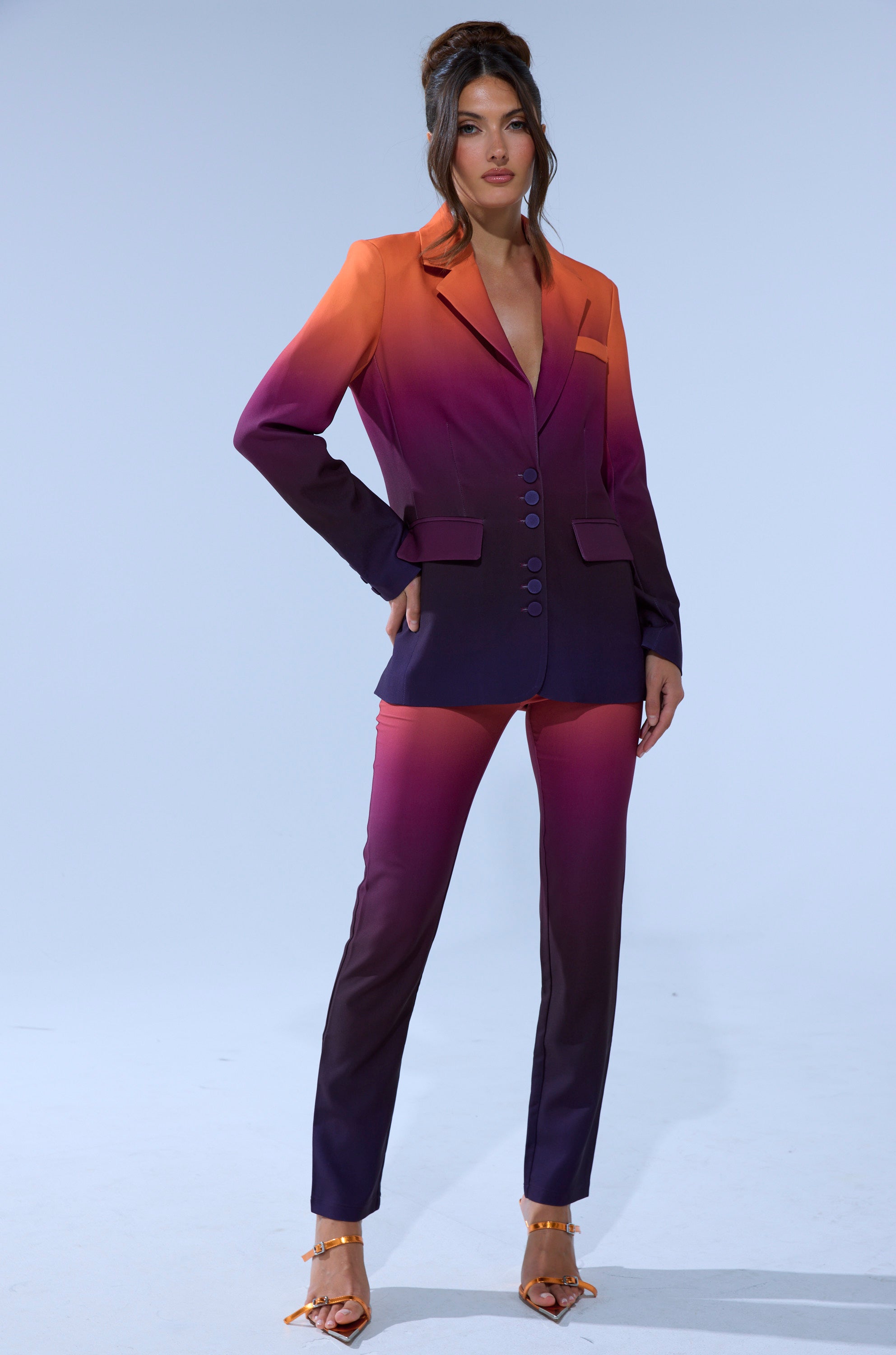 A woman stands confidently in the RISE AND SHINE OMBRE BLAZER and matching pantsuit, featuring orange, pink, and purple hues with strappy heels, against a light gray background.