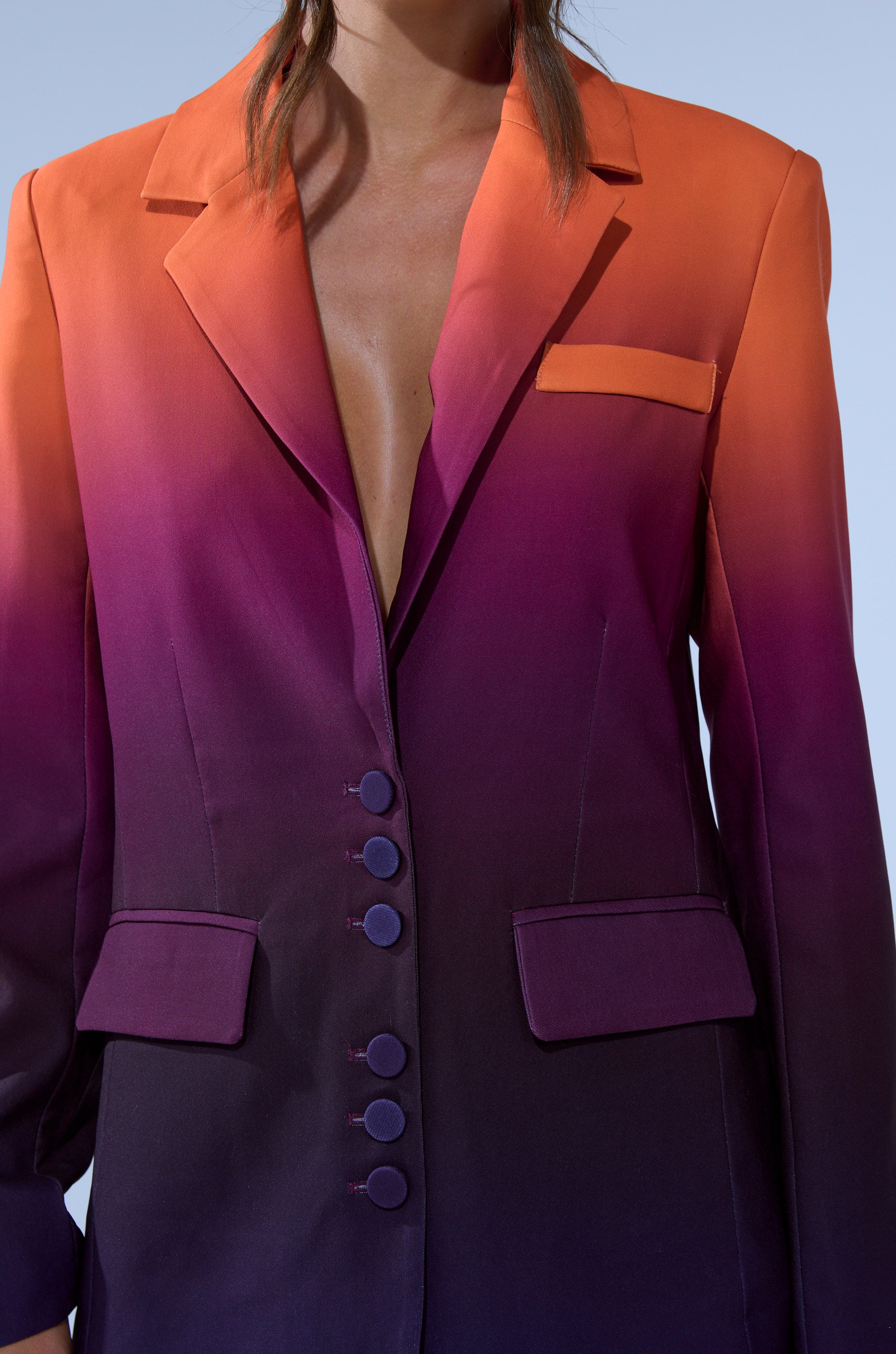 A person wears the RISE AND SHINE OMBRE BLAZER, featuring a gradient from orange shoulders to deep purple hem, a plunging neckline, and five front buttons for a bold sunset-inspired look.