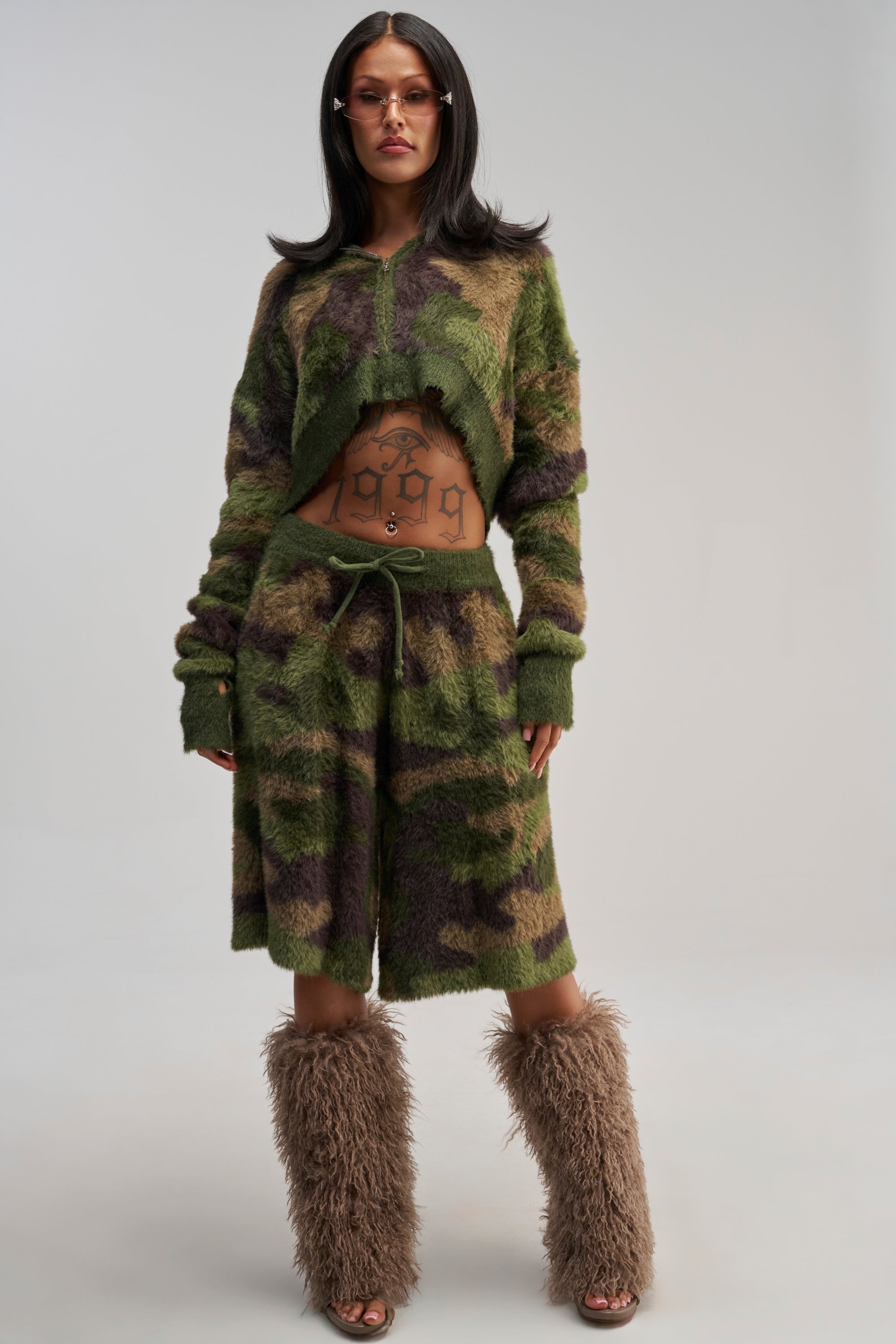 Wearing the GO WILD FUZZY BERMUDA SHORT, a woman confidently poses against a plain background, pairing the green and brown fuzzy shorts with a matching long sleeve top, furry brown leg warmers, sunglasses, and revealing midriff tattoos for an eye-catching streetwear look.
