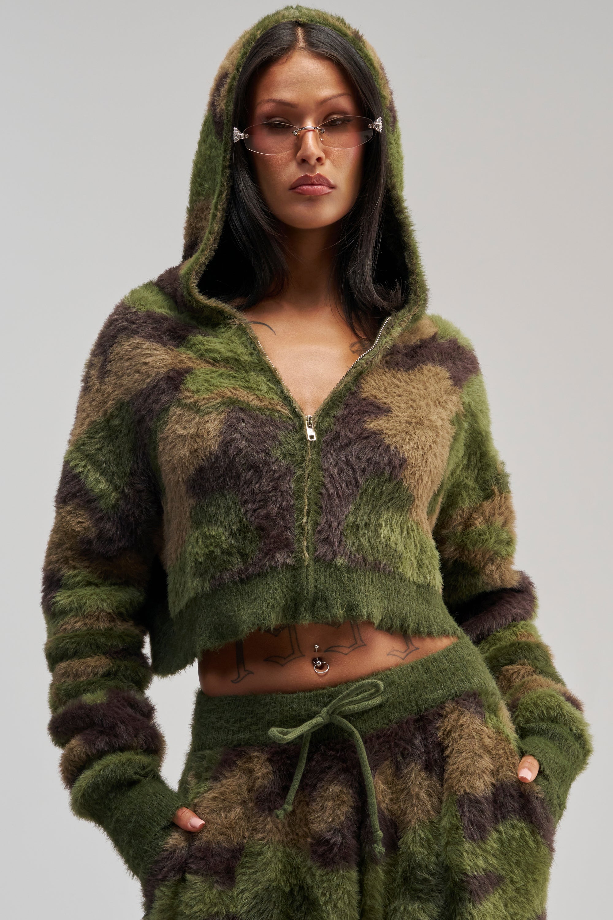 Wearing the GO WILD FUZZY SWEATSHIRT, a woman poses confidently in a cozy green and brown camo zip-up with matching pants, embodying streetwear loungewear style and accessorizing with small, narrow sunglasses.