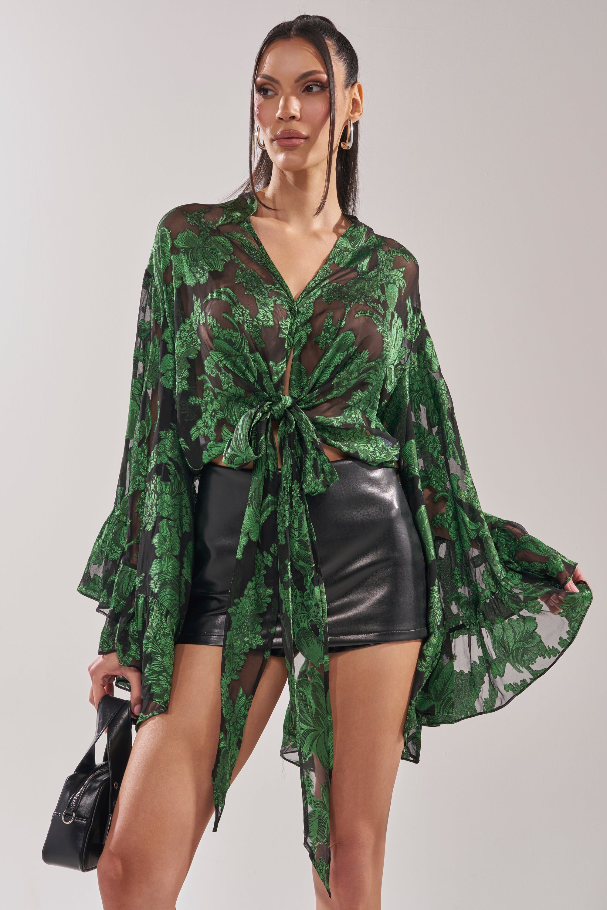 A woman with long, straight hair stands against a light background wearing the IN THE FEELS LACE BLOUSE, featuring a sheer green botanical pattern and tied at the waist, styled with black leather shorts and a small black handbag. 0