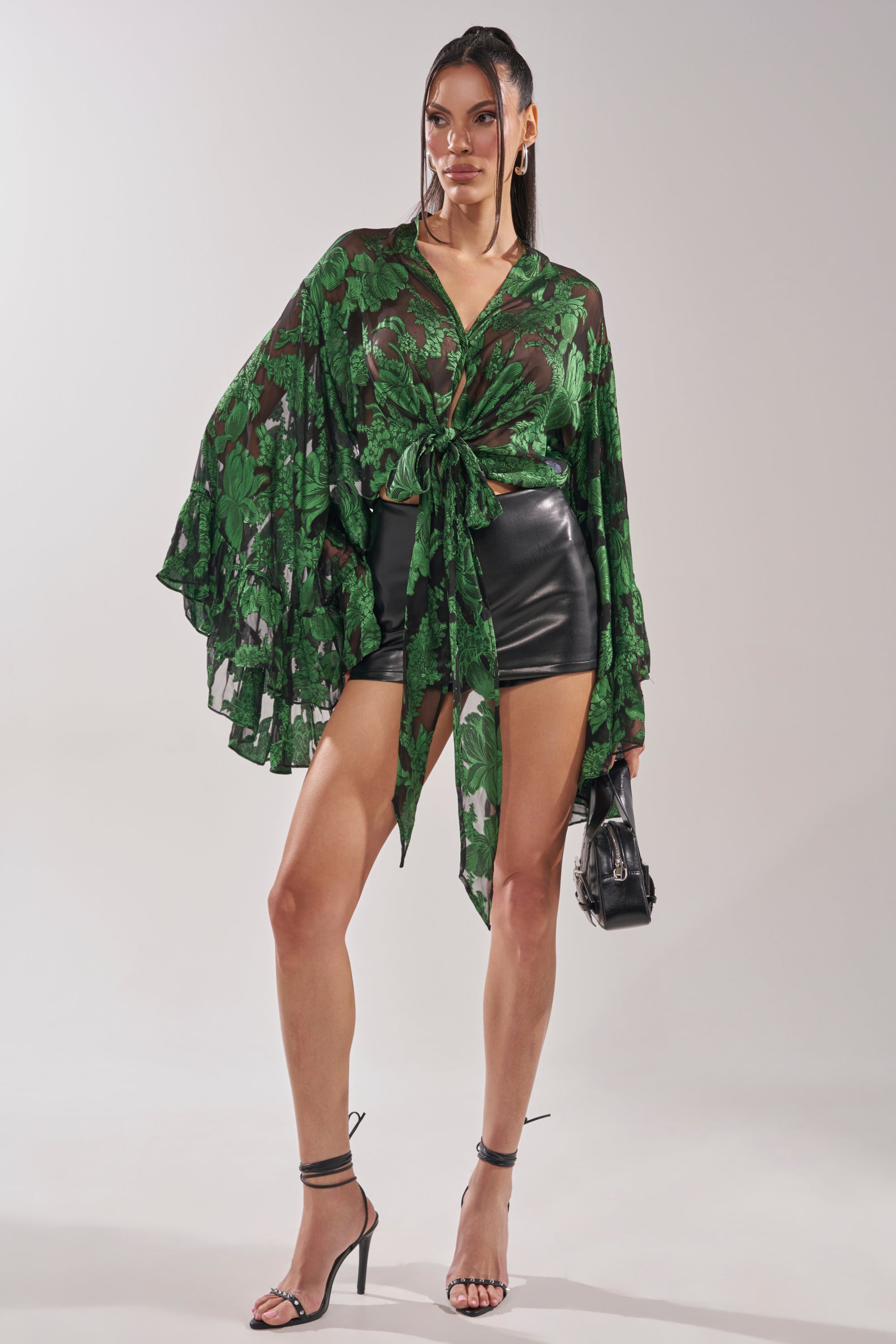 A woman poses in the IN THE FEELS LACE BLOUSE—a sheer green floral cropped top with wide sleeves and waist tie—styled with black leather shorts, high heels, and a small black handbag against a plain background. 9