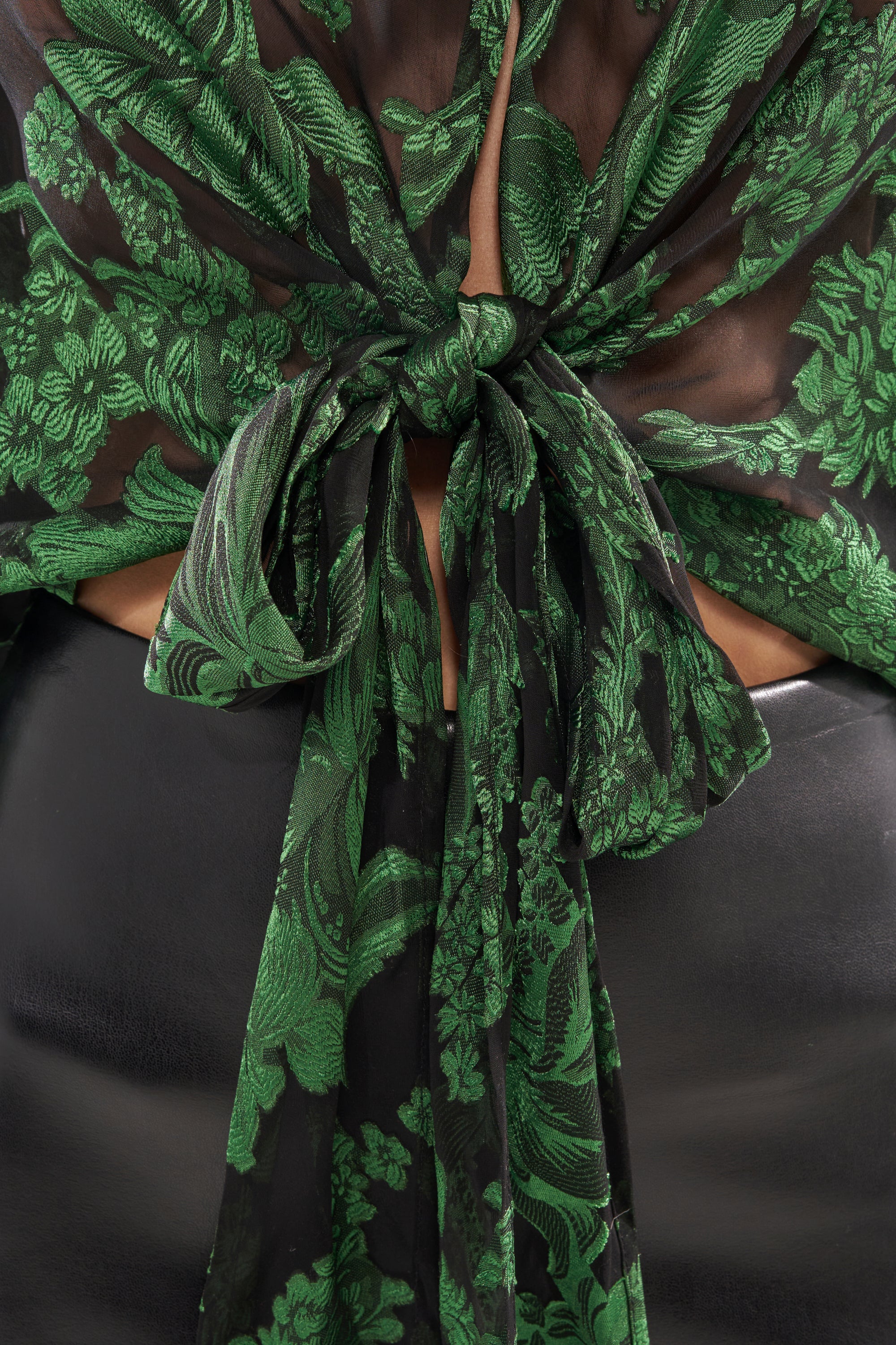 Close-up of the IN THE FEELS LACE BLOUSE featuring sheer floral fabric, a back bow tie, green embroidery, and paired with shiny black bottoms. 10