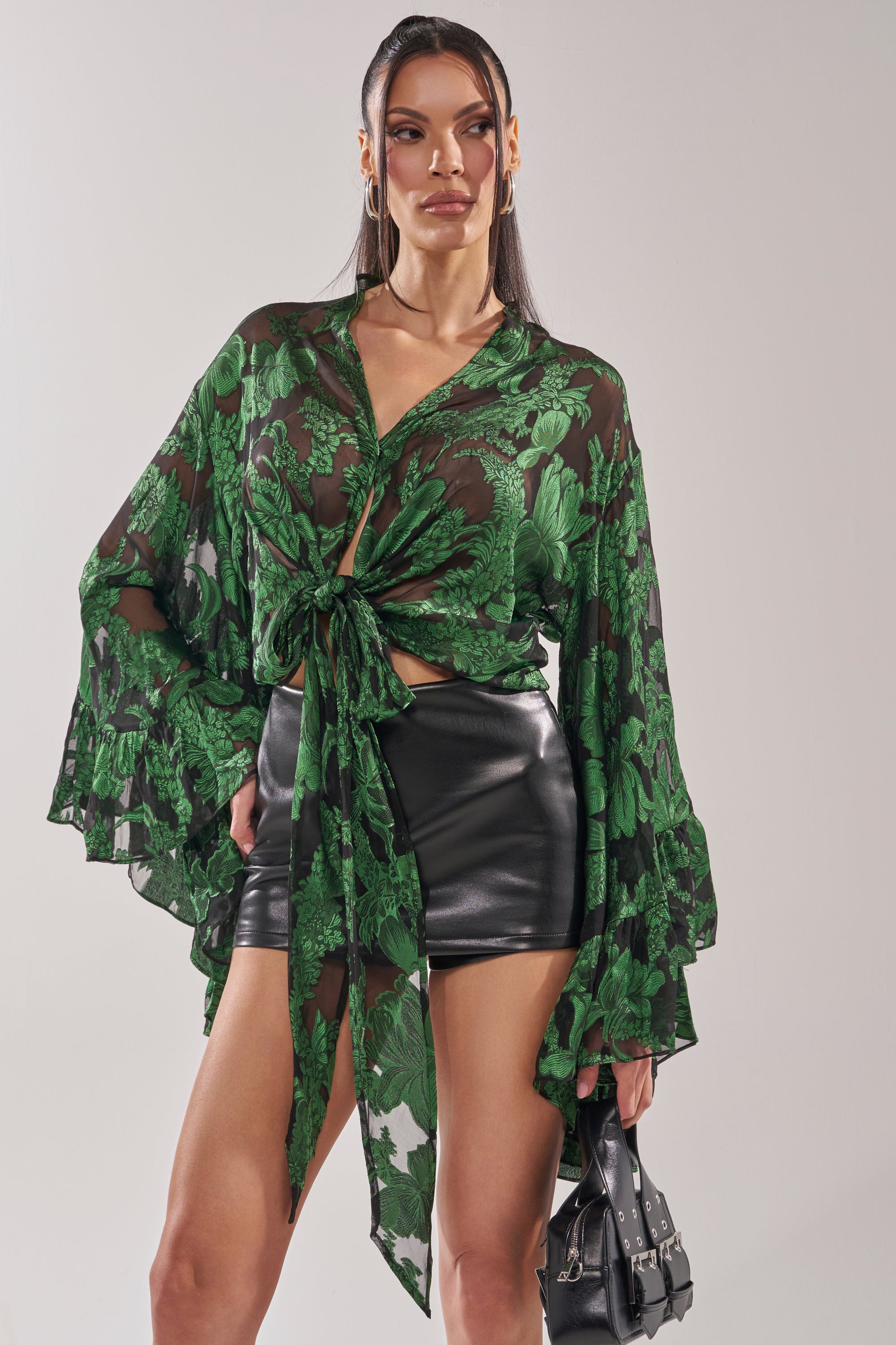 A woman poses in the IN THE FEELS LACE BLOUSE, styled with a waist knot and wide sleeves, paired with a black leather mini skirt, hoop earrings, and a small black handbag. 1