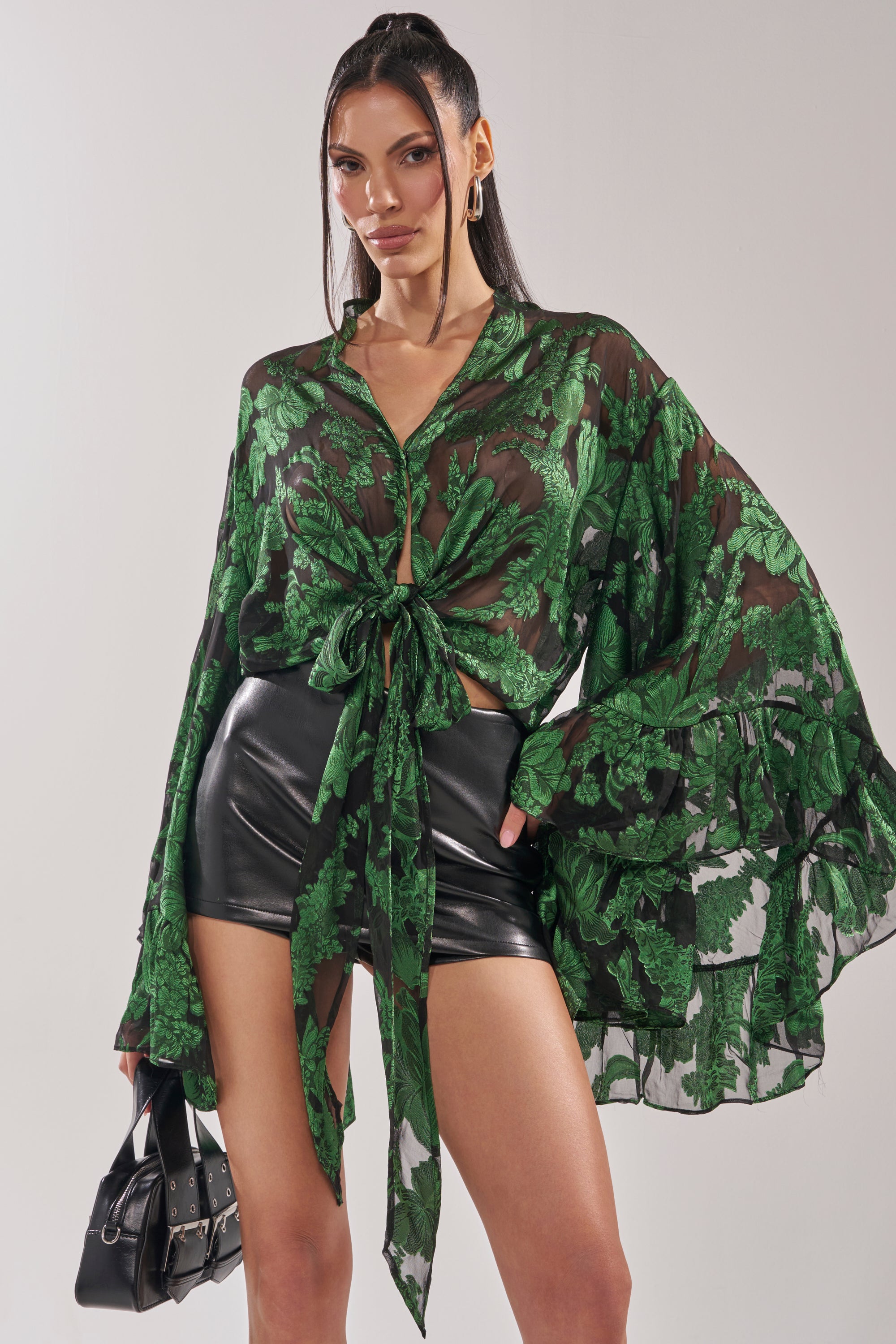 A woman exudes confidence in the IN THE FEELS LACE BLOUSE, featuring a sheer green botanical pattern and wide sleeves, paired with black high-waisted leather shorts. Her sleek hair and black handbag complete this chic look. 2