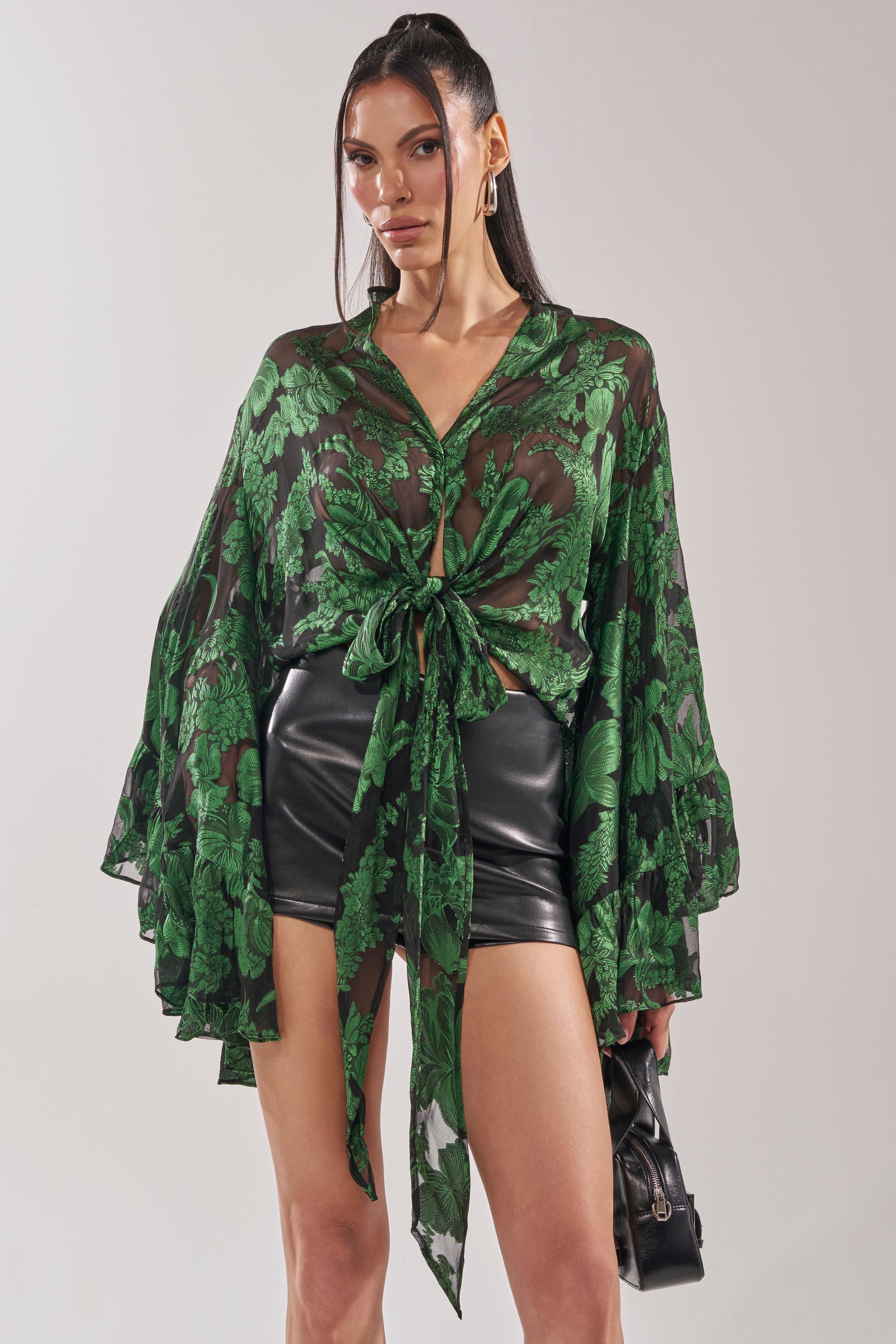 A woman confidently poses in high-waisted black leather shorts and the IN THE FEELS LACE BLOUSE—a sheer green top with a lace botanical pattern and flared sleeves—while holding a small black handbag. 3