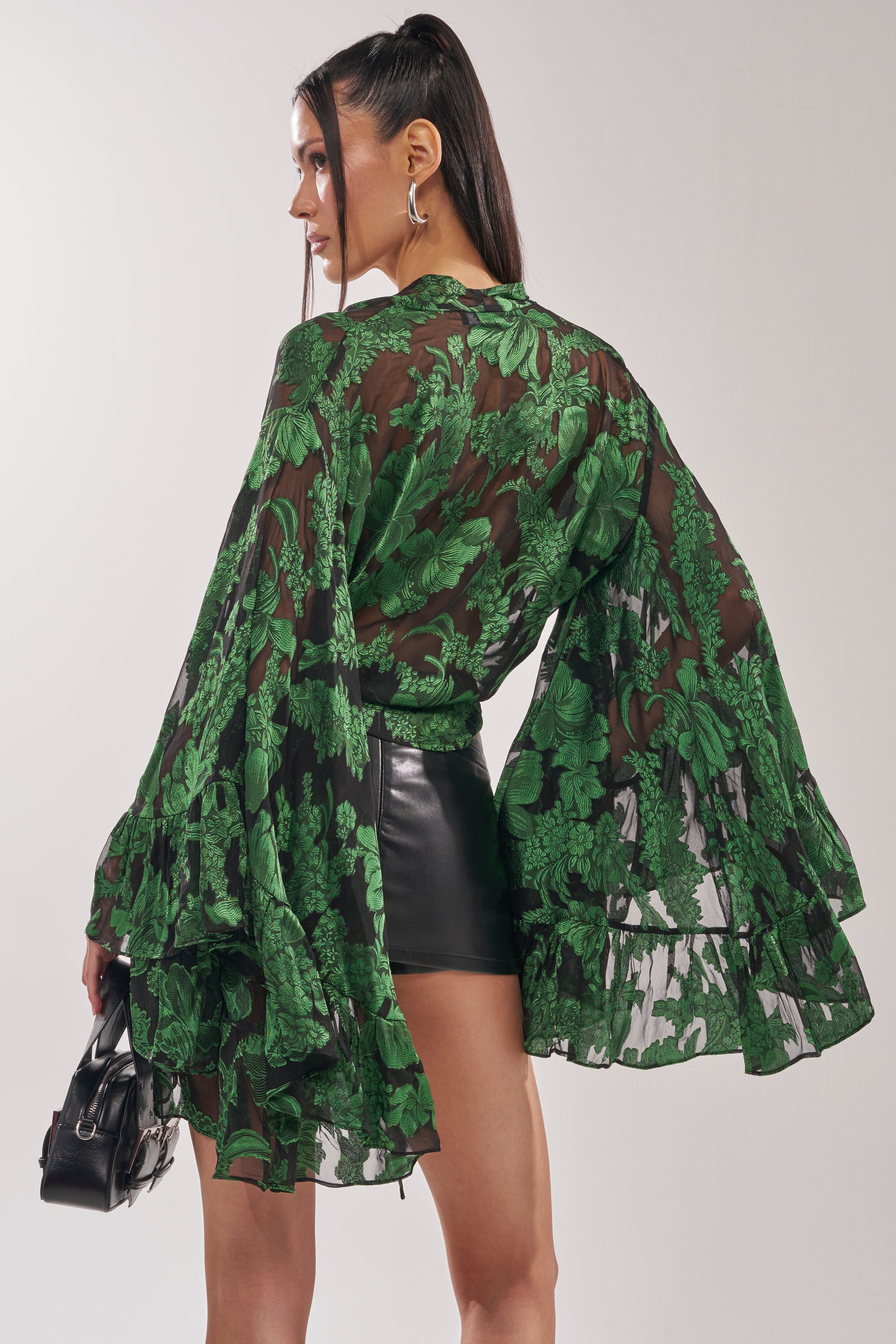 Woman faces away, wearing the IN THE FEELS LACE BLOUSE with sheer floral fabric and ruffled wide sleeves, paired with black faux leather shorts. She carries a small black handbag; her long dark hair is in a high ponytail. 5