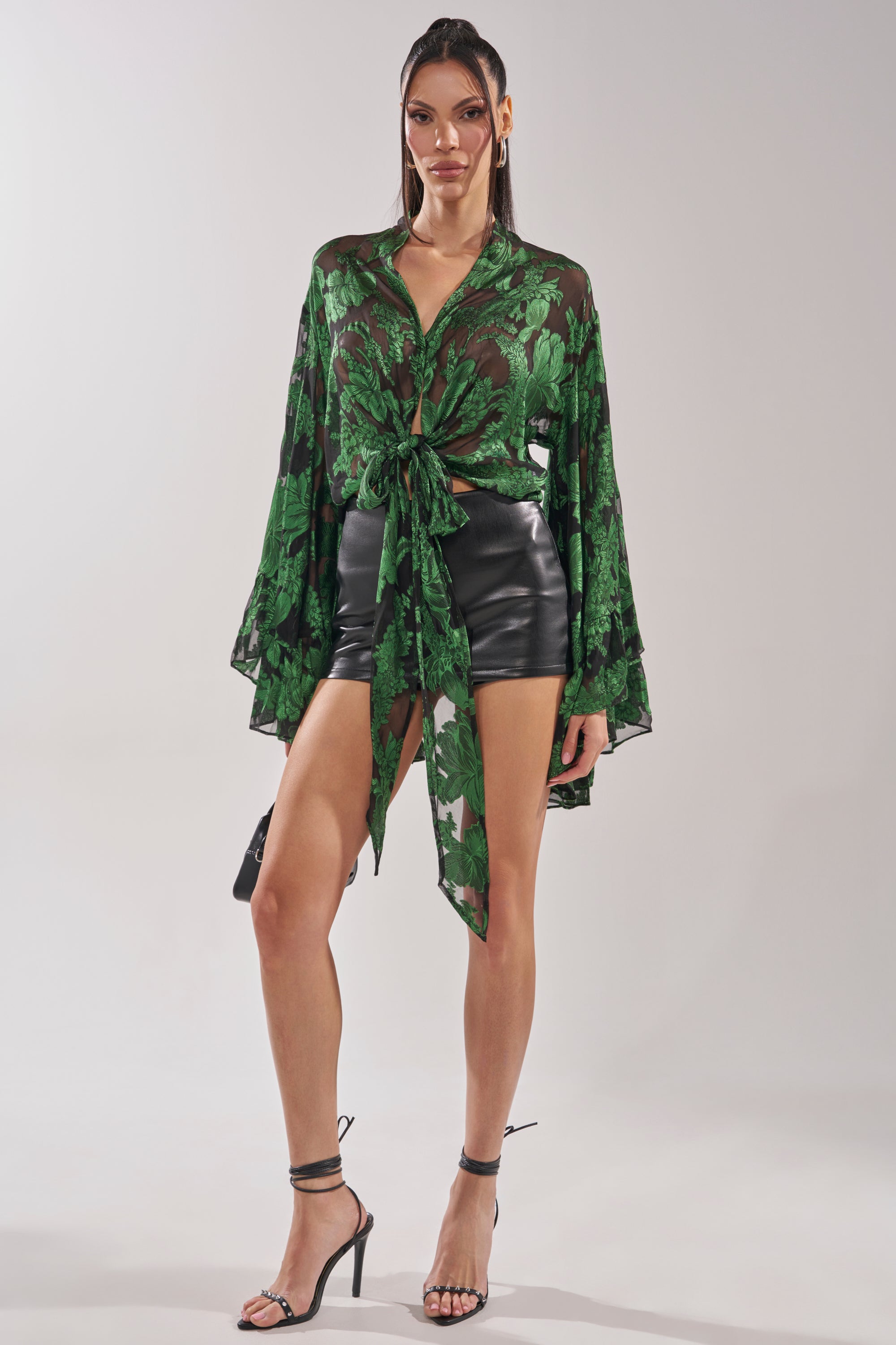 A woman poses against a plain background in the IN THE FEELS LACE BLOUSE, a sheer green and black floral crop top tied at the waist, styled with black high-waisted leather shorts and black high-heeled sandals. 7