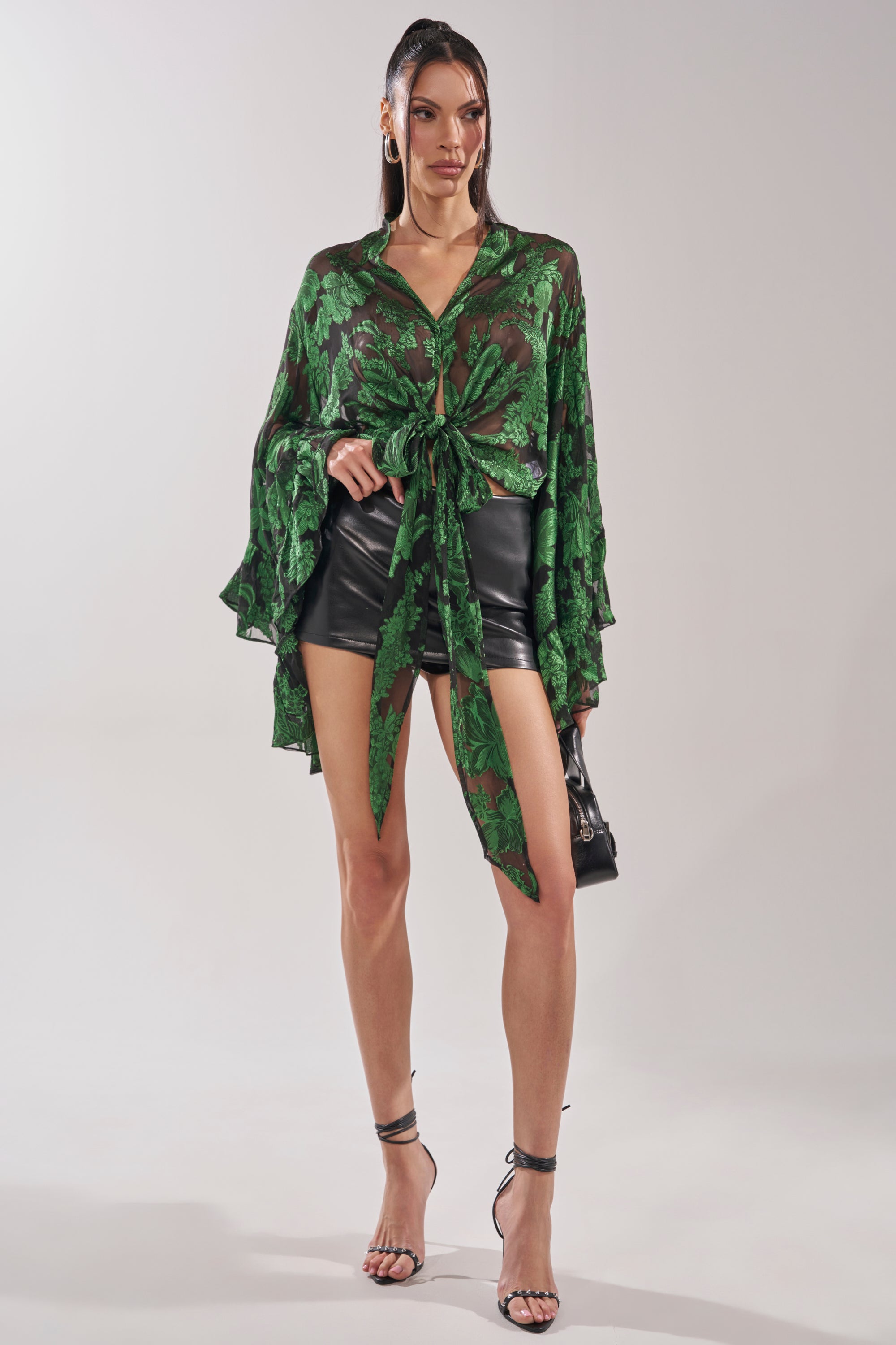 A woman poses against a plain background in the IN THE FEELS LACE BLOUSE with wide sleeves, paired with black leather shorts, high-heeled sandals, and a small black handbag. 8