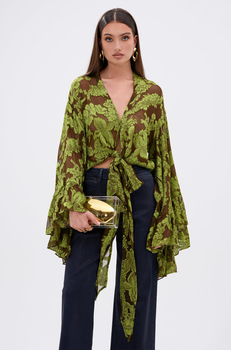 A woman wearing the BUTTERFLY EFFECT BLOUSE IN GREEN with floral burnout, tiered bell sleeves, dark wide-leg pants, gold hoop earrings, and a gold clutch stands against a plain white background.