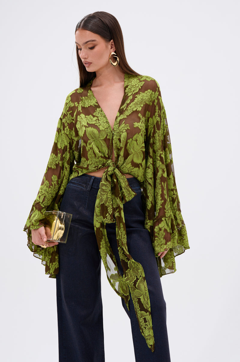 A woman wears the BUTTERFLY EFFECT BLOUSE IN GREEN, a chartreuse floral burnout with dramatic tiered bell sleeves, paired with dark wide-leg pants. She's styled with a gold clutch and bold gold hoops, looking down to the side.
