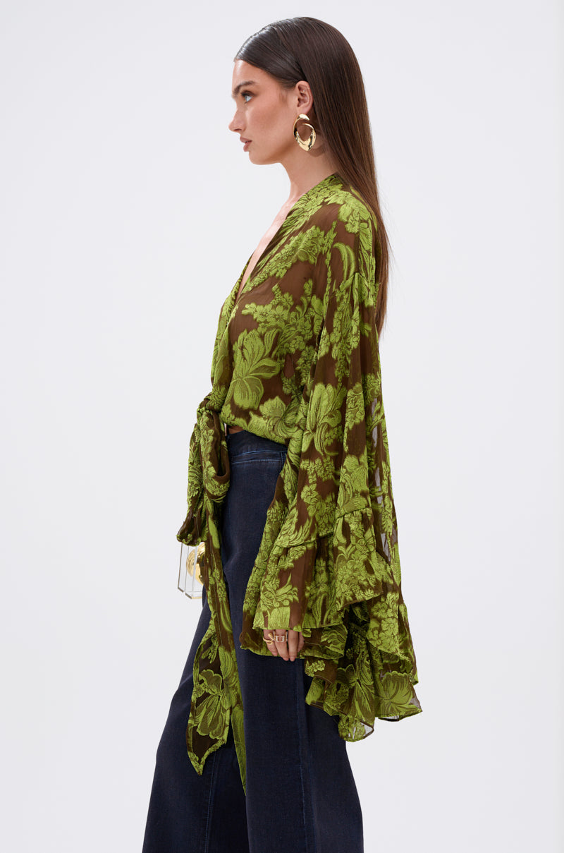A woman stands in profile, wearing the BUTTERFLY EFFECT BLOUSE IN GREEN with floral burnout fabric, exaggerated tiered bell sleeves, and dark high-waisted jeans. She has long straight hair, large hoop earrings, and a neutral background.