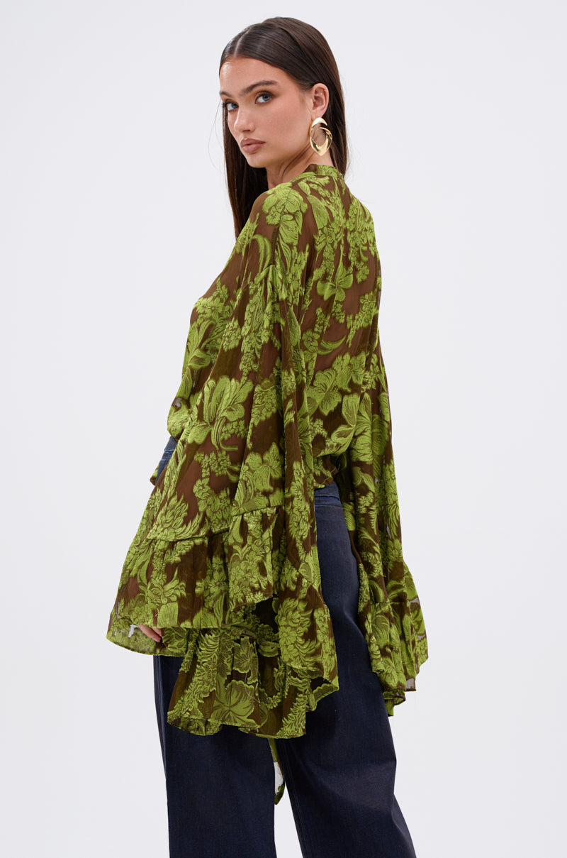 A woman with long brown hair, wearing large gold hoop earrings and the Butterfly Effect Blouse in green with exaggerated tiered bell sleeves, stands sideways in dark wide-leg pants against a plain white background.