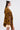 A woman in the ADORE YOU TIE FRONT BLOUSE with golden floral print and bell sleeves pairs it with brown leather shorts, a beige clutch, gold earrings, and layered necklaces while standing in profile against a white background.