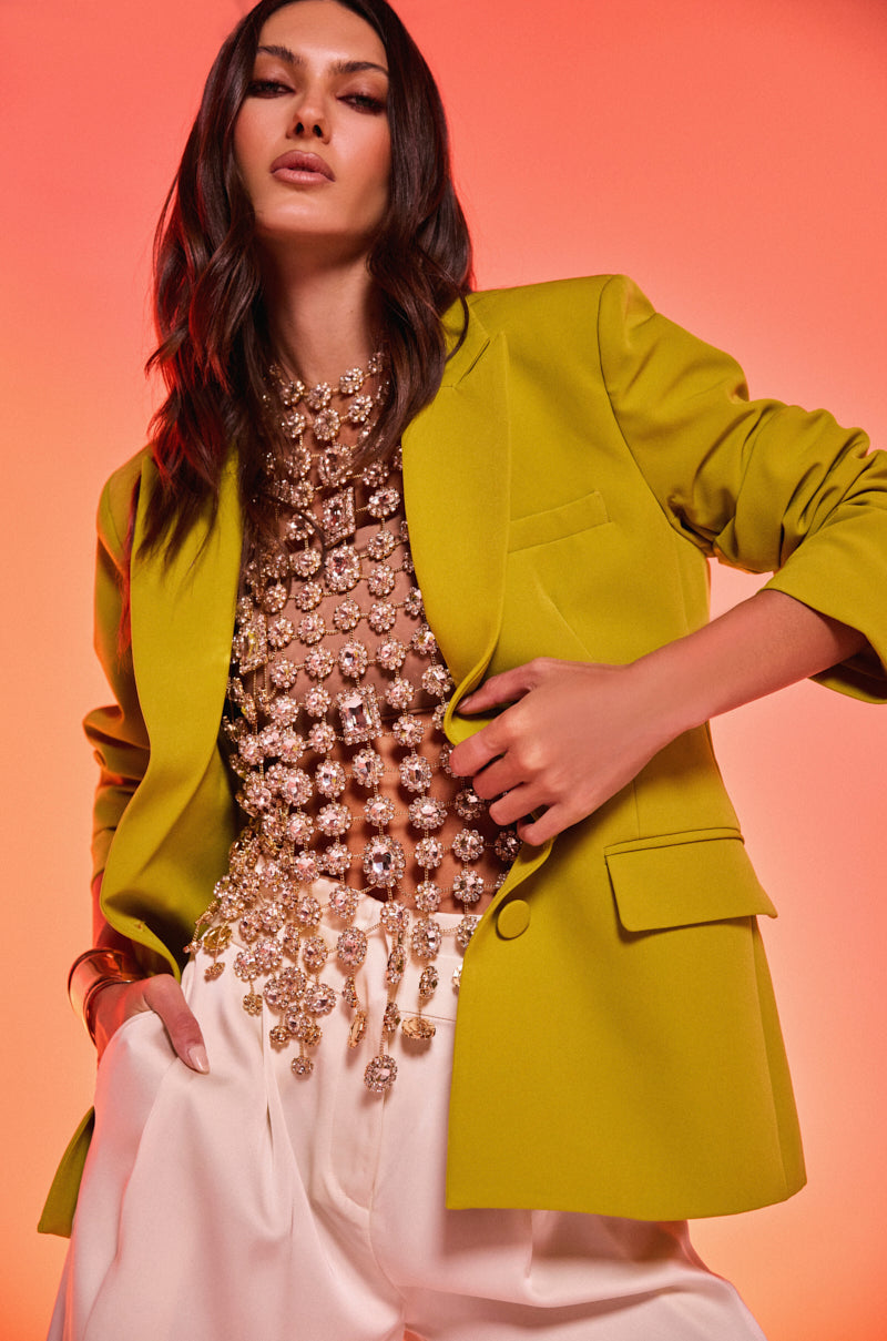 A woman poses confidently against a warm orange backdrop, wearing the IRIS BLAZER IN CHARTREUSE over a jeweled sheer top and high-waisted white pants, with one hand in her pocket and the other on her waist.