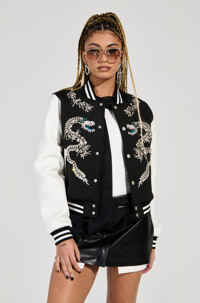 Wearing sunglasses and the eye-catching DRAGONETTE EMBELLISHED BOMBER adorned with dragon embroidery and shimmering accents, a person with long braided hair teams it with a leather mini skirt. They pose confidently against a neutral background, effortlessly radiating coolness.
