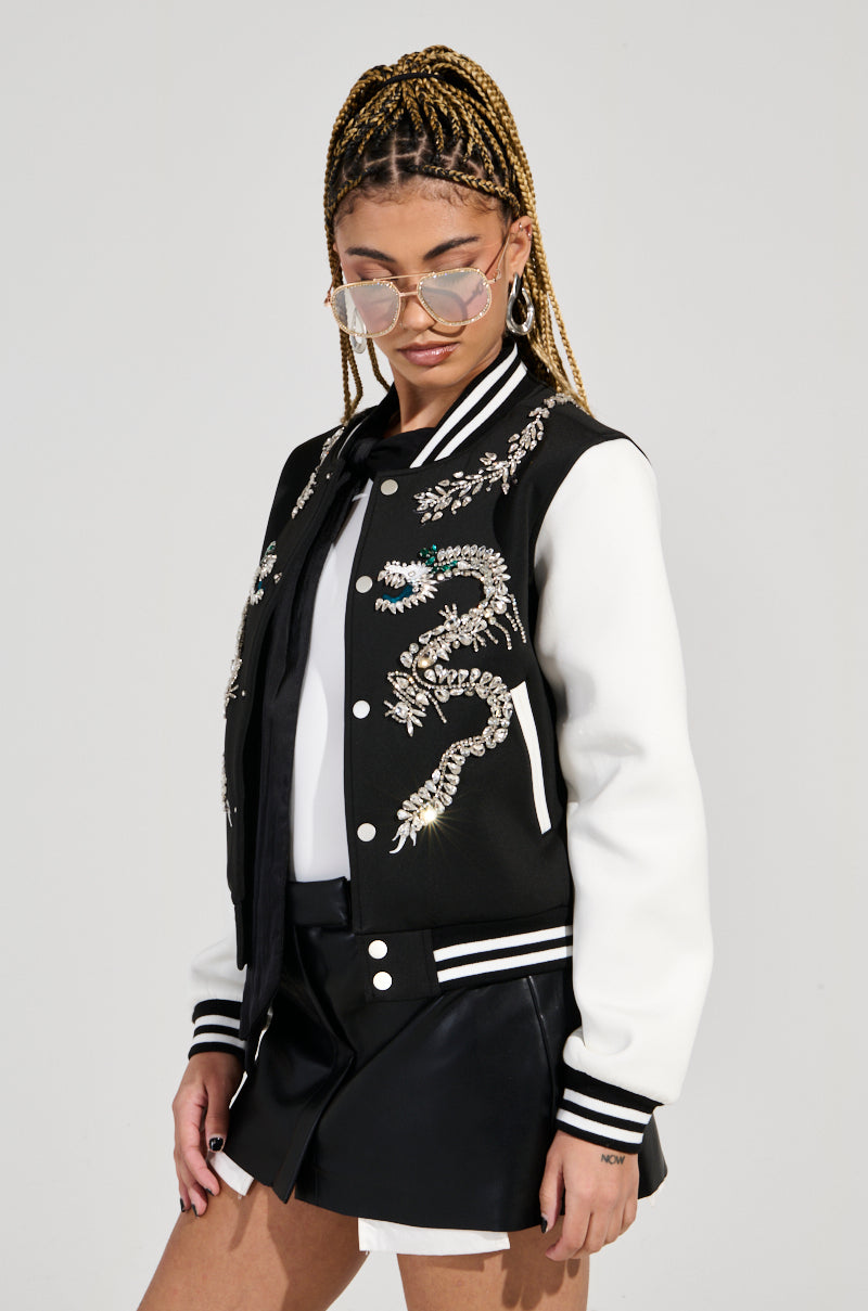 A person dressed confidently in a chic black and white DRAGONETTE EMBELLISHED BOMBER, adorned with intricate dragon motifs, exudes style. They complement their look with braided hair, sunglasses, a crisp white shirt, and a sleek black skirt against a plain backdrop that enhances their fashion-forward ensemble.