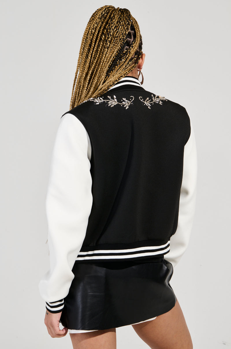 Rear view of a person with long braided hair wearing the DRAGONETTE EMBELLISHED BOMBER, a black and white varsity jacket adorned with dragon embroidery. They are also wearing a black mini skirt.