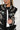 Dressed in a DRAGONETTE EMBELLISHED BOMBER adorned with detailed dragon embroidery, rhinestones, and faux emeralds, the individual complements their long braided hair with round sunglasses and hoop earrings.