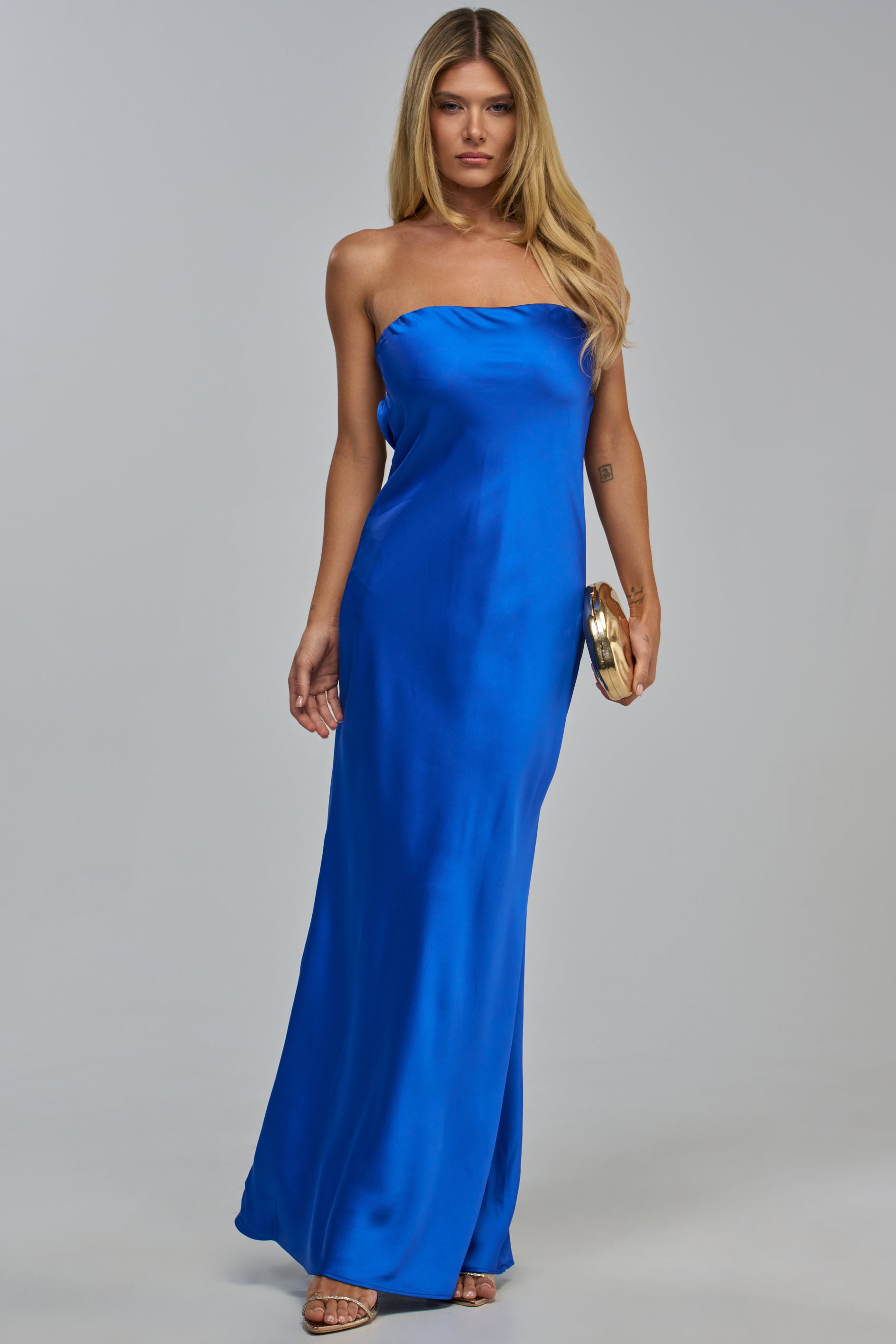 A woman wearing a royal blue STILL YOURS SATIN STRAPLESS MAXI DRESS, styled as a floor-length strapless gown and holding a gold clutch.