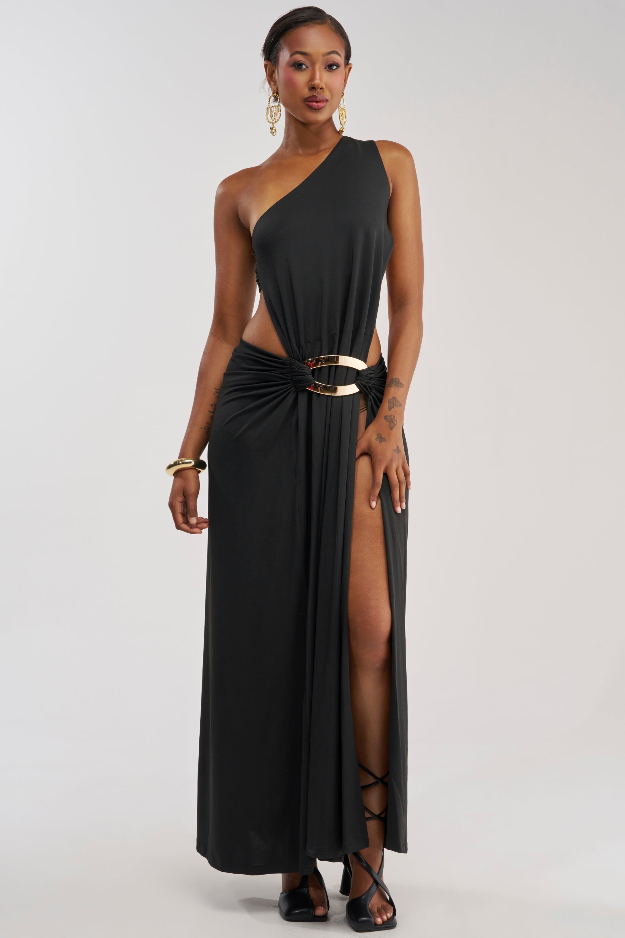 A woman showcases the HEARTBREAKER ONE SHOULDER MAXI DRESS IN BLACK—a striking maxi dress featuring a one-shoulder design, high slit, and waist cutout. She completes the look with gold accessories, black sandals, and sleekly styled hair against a plain, light backdrop.