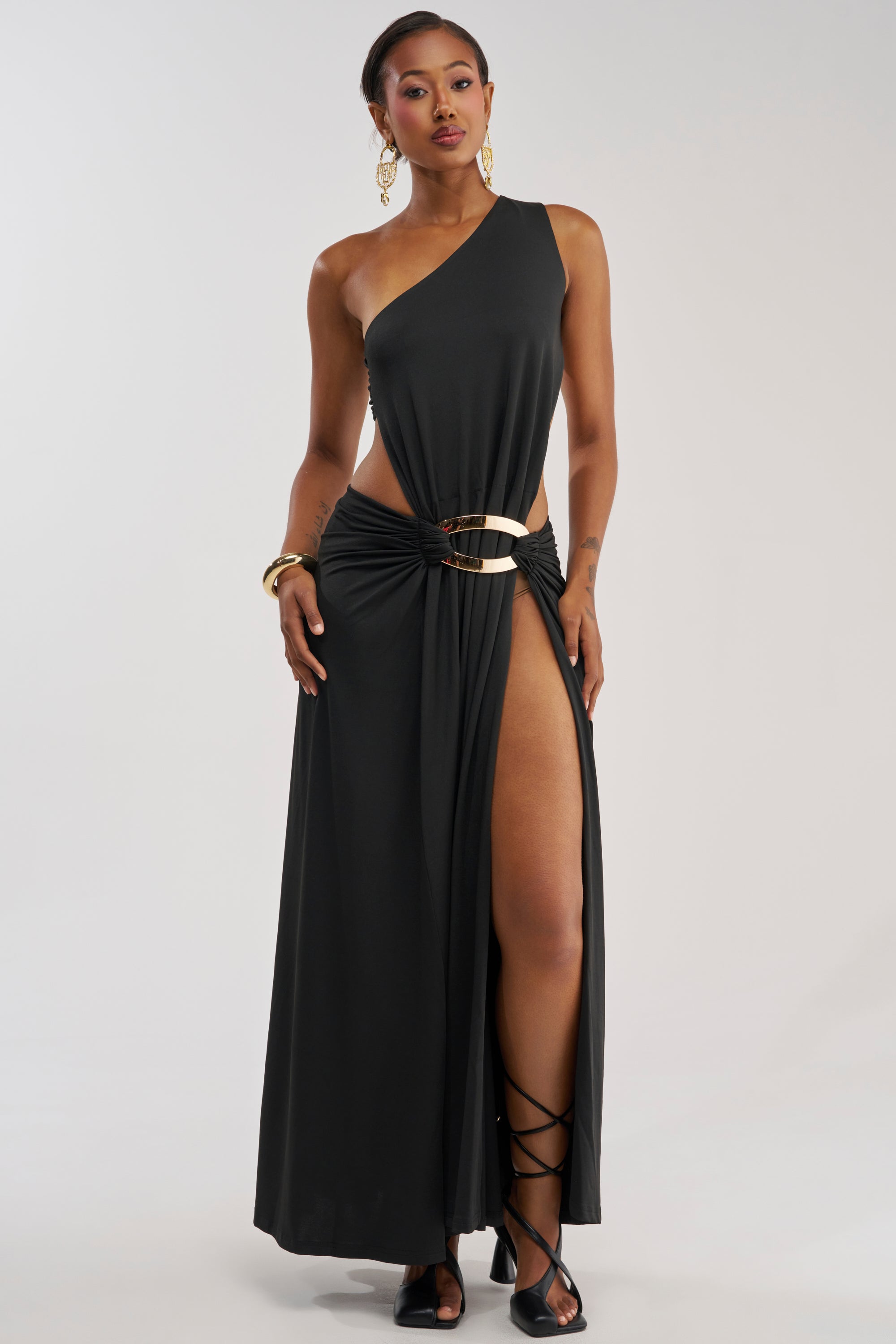 A woman stands against a plain light background wearing the HEARTBREAKER ONE SHOULDER MAXI DRESS IN BLACK, featuring a gold ring detail at the waist and a high slit, styled with gladiator sandals, gold jewelry, and hoop earrings.