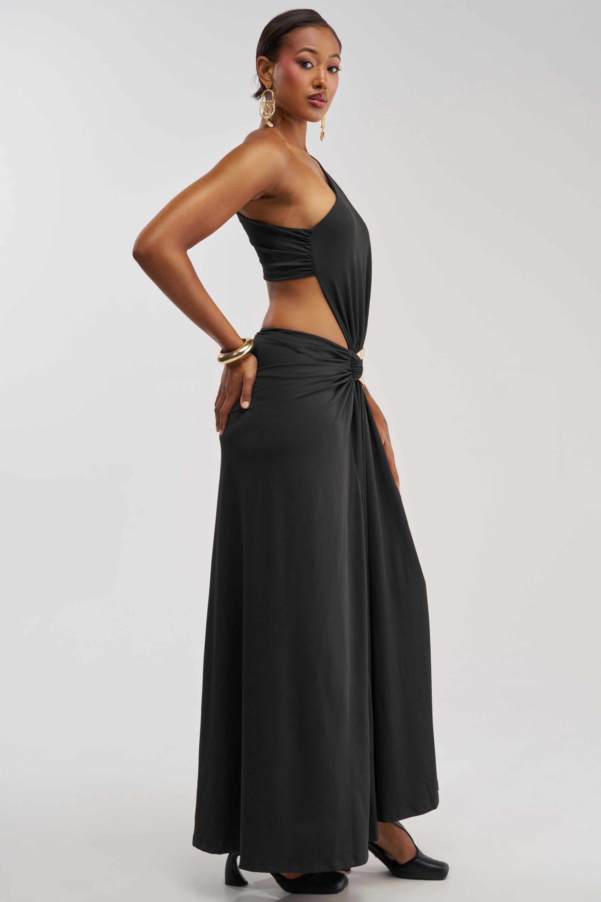 A woman poses in the HEARTBREAKER ONE SHOULDER MAXI DRESS IN BLACK—a stylish black cutout maxi dress with asymmetrical, one-shoulder detailing—paired with gold earrings, black shoes, and gold bangles against a plain light background.
