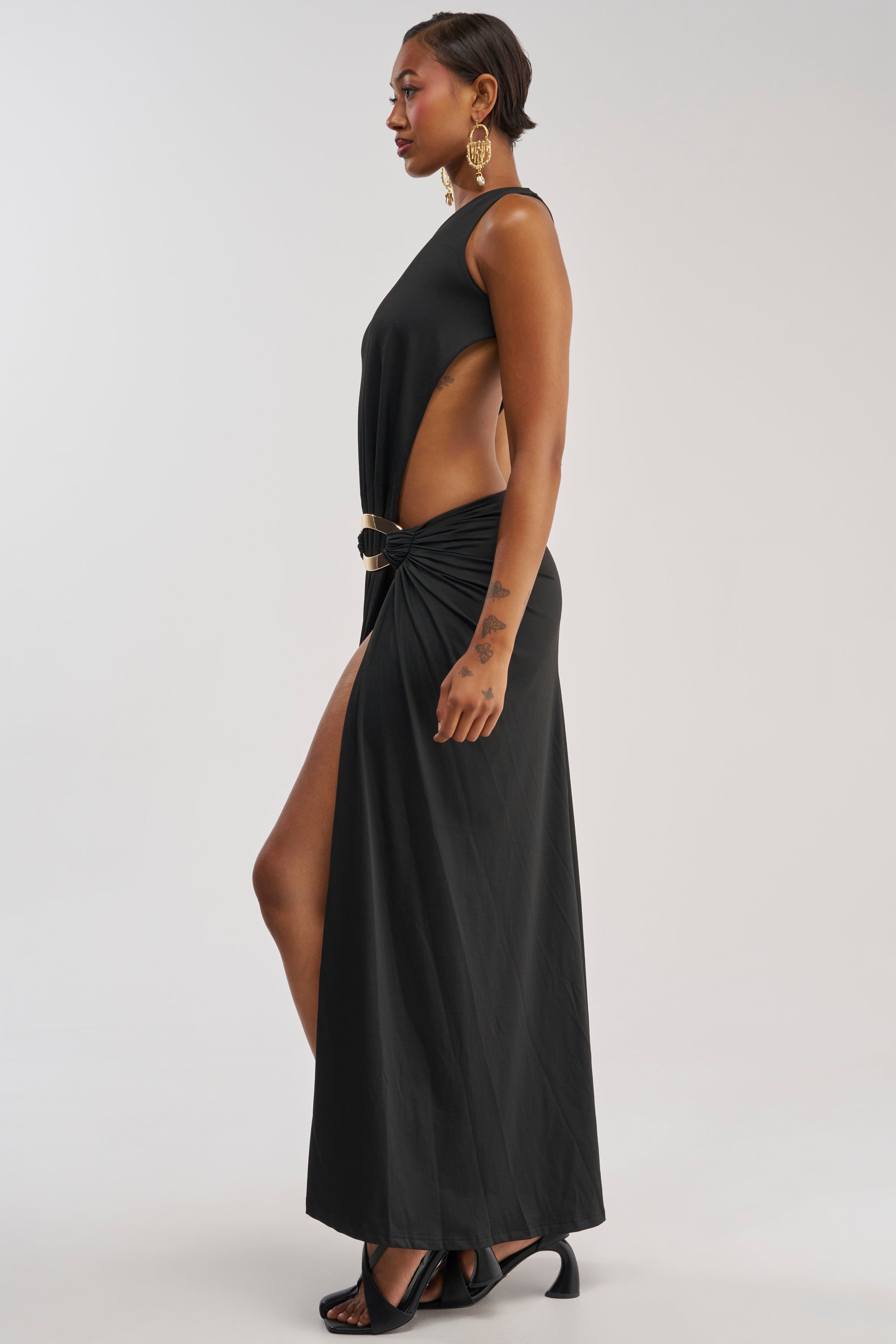 A woman wearing the HEARTBREAKER ONE SHOULDER MAXI DRESS IN BLACK, styled with gold earrings and black heels, stands sideways against a plain light background.