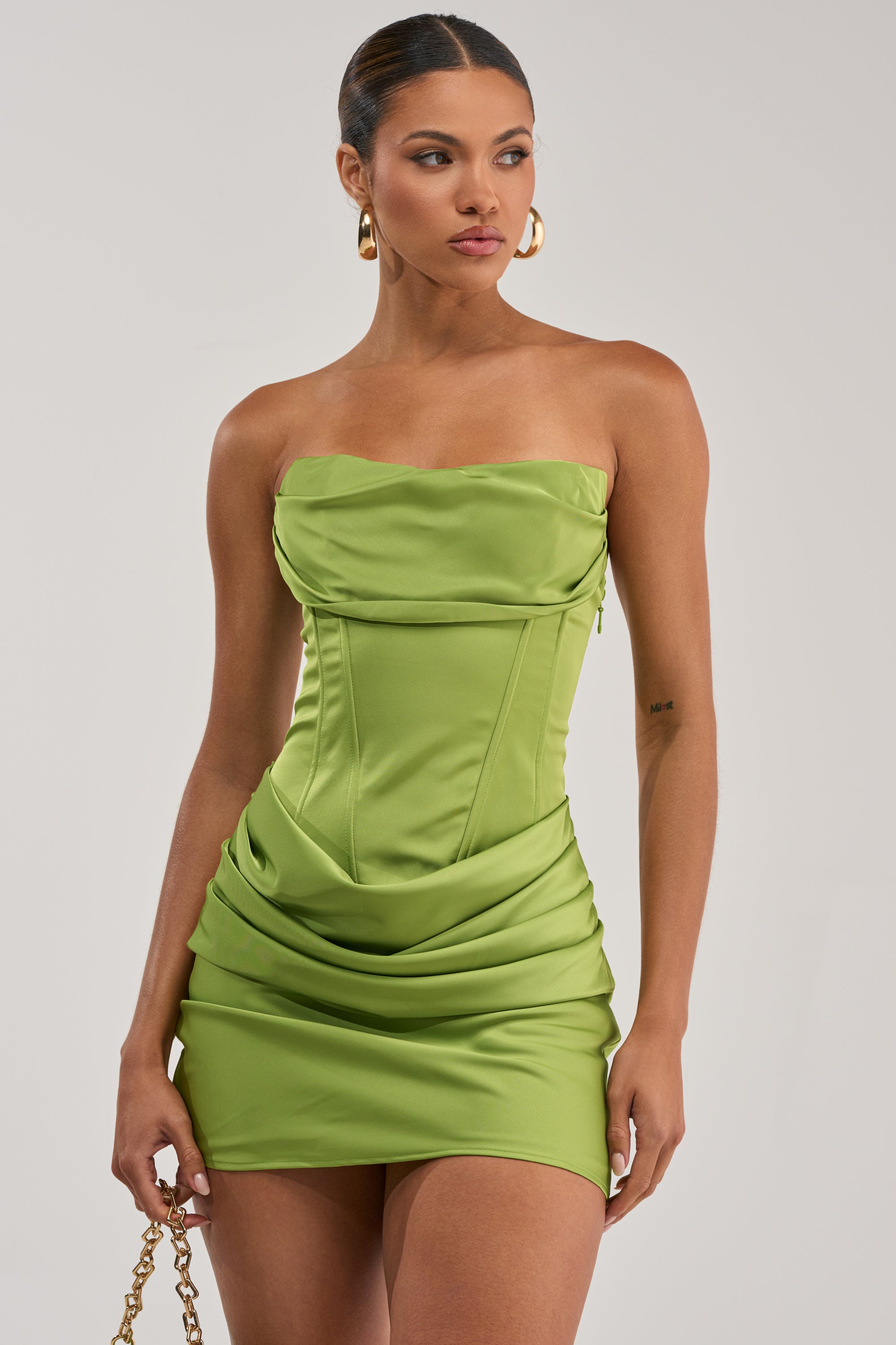 A woman models the NO COMPETITION STRAPLESS SATIN MINI DRESS IN CHARTREUSE— a fitted mini with draped details and structured bodice—styled with gold hoop earrings and a gold chain strap, posing against a plain light background.