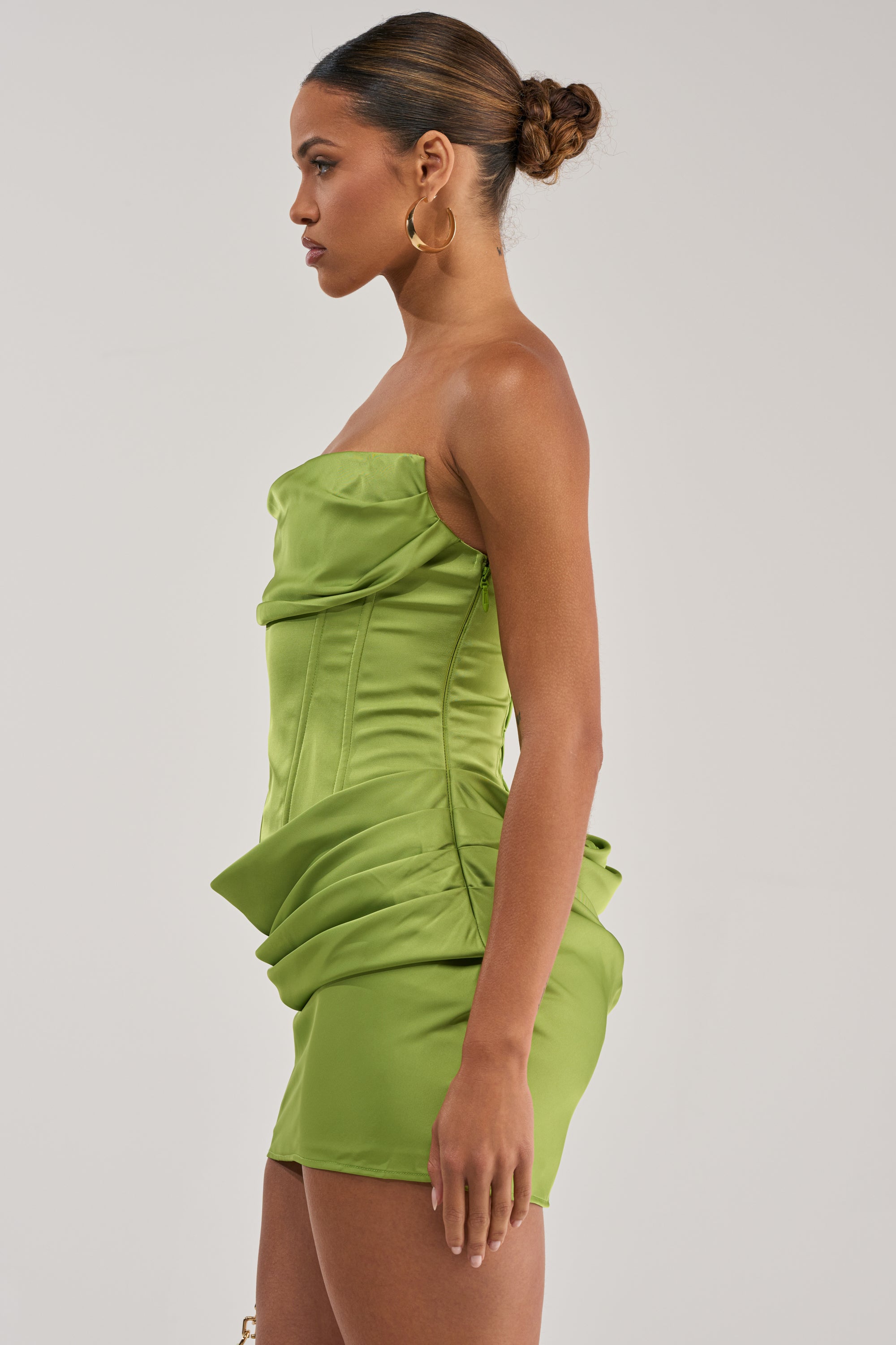 A woman with her hair in a bun models the NO COMPETITION Strapless Satin Mini Dress in chartreuse, featuring a structured bodice and draped details, standing in profile against a plain light background.