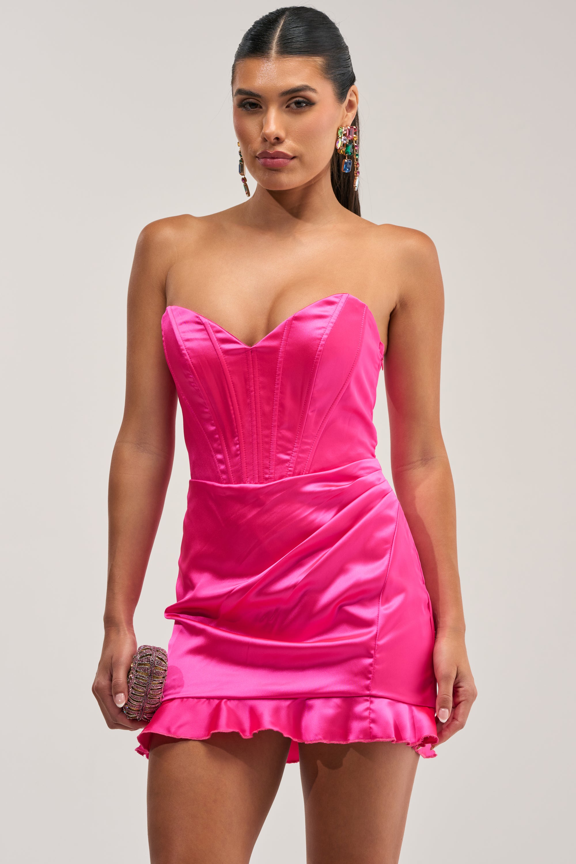 A woman wearing THE TRUTH ABOUT LOVE SATIN MINI DRESS IN PINK, a strapless hot pink mini dress featuring a sweetheart neckline and ruffled hem, holds a small clutch and completes the look with colorful earrings.