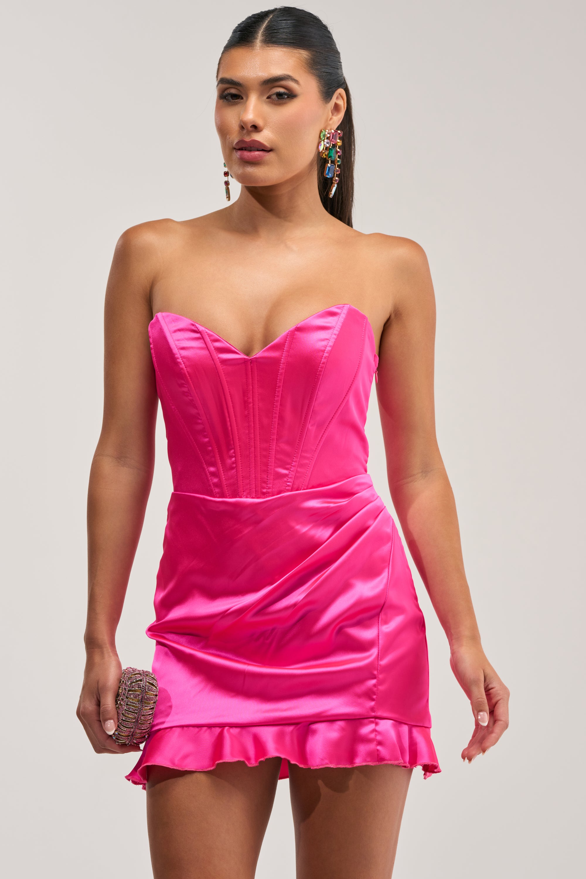 A woman wears THE TRUTH ABOUT LOVE SATIN MINI DRESS IN PINK, featuring a strapless design, sweetheart neckline, fitted bodice, and ruffled hem. She accessorizes with a small sparkly clutch and colorful earrings, her long hair styled beautifully as she strikes a confident pose.