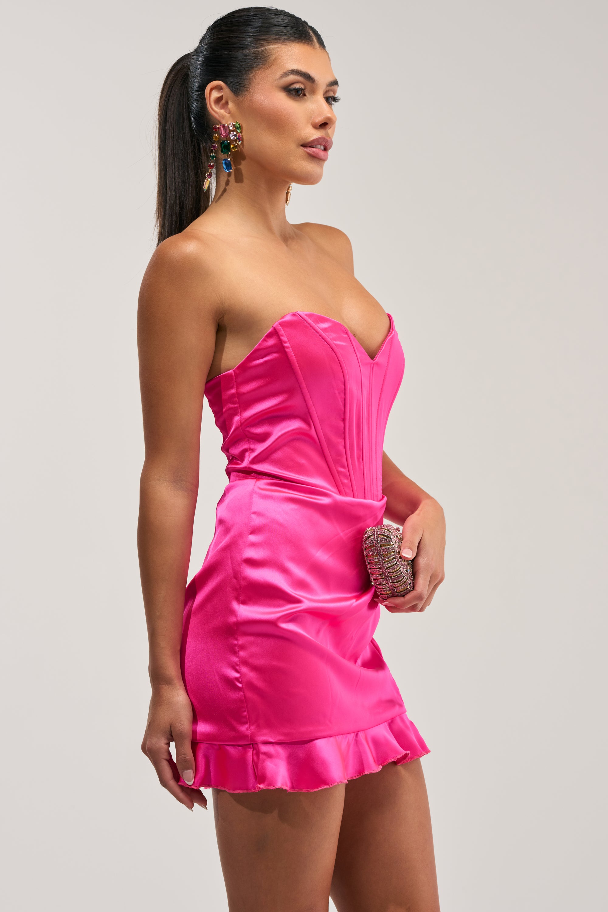 Wearing THE TRUTH ABOUT LOVE Satin Mini Dress in Pink, a woman stands against a plain light background. The hot pink strapless mini dress features a ruffled hem; she completes the look with a small clutch and colorful earrings.