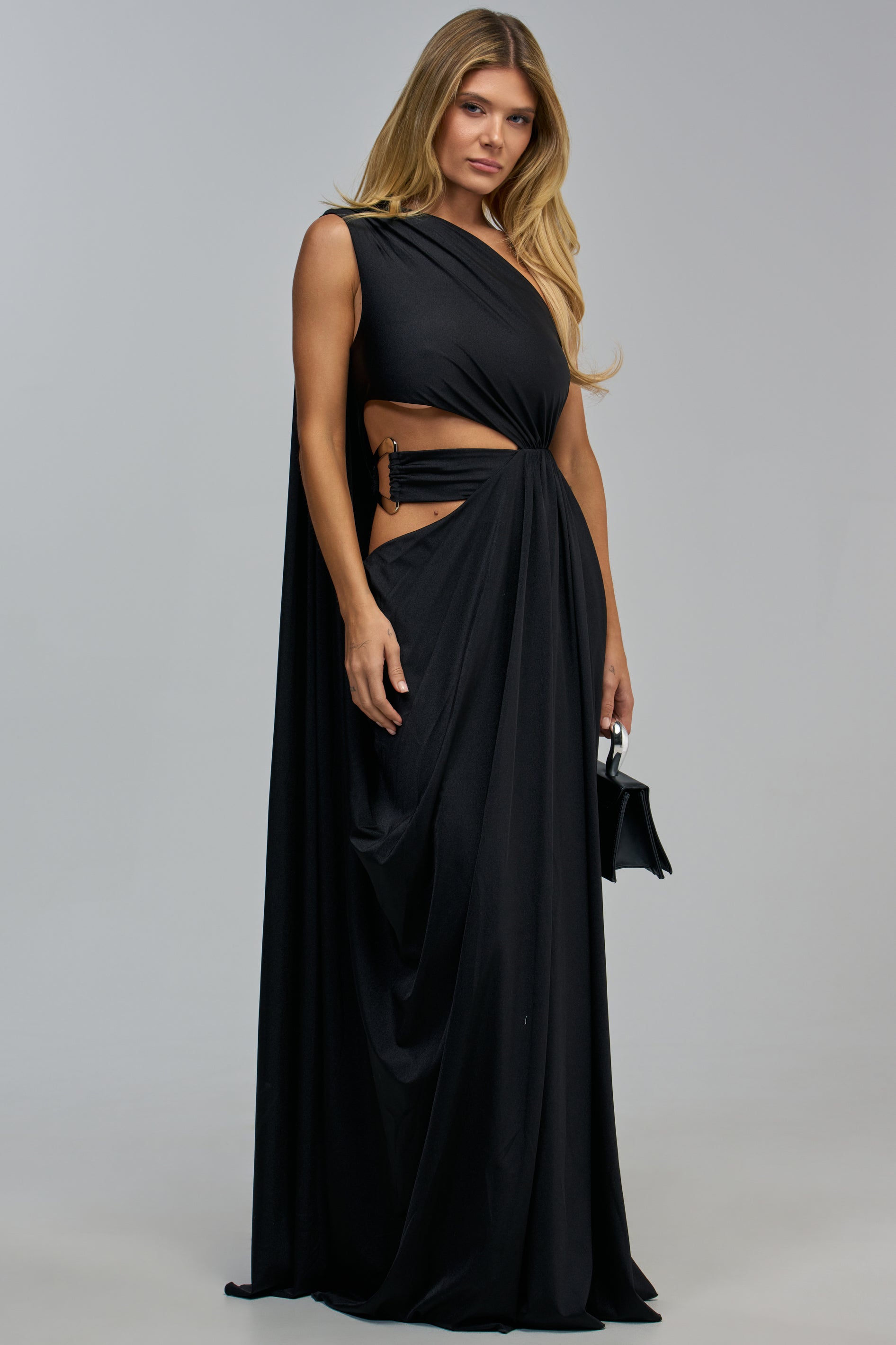 A woman wears a black DON’T GET IT TWISTED CUT-OUT MAXI DRESS featuring a one-shoulder neckline, side cut-outs, and a gold ring detail at the waist, paired with a small black handbag.