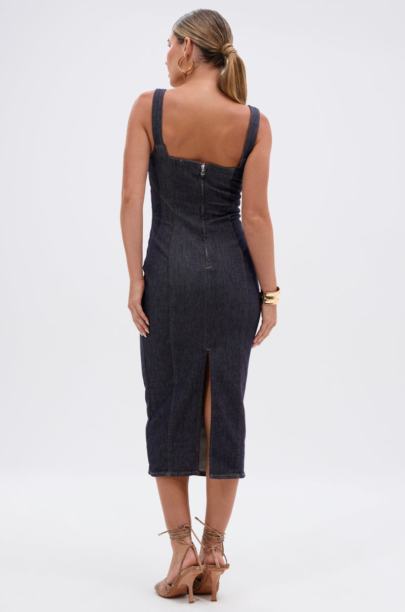 A woman stands facing away in the DAY DREAMIN DENIM MIDI DRESS, a dark-wash, body-hugging silhouette with a back zipper and center slit. Her hair is tied back, accessorized with gold bracelets and strappy heels against a plain white background.