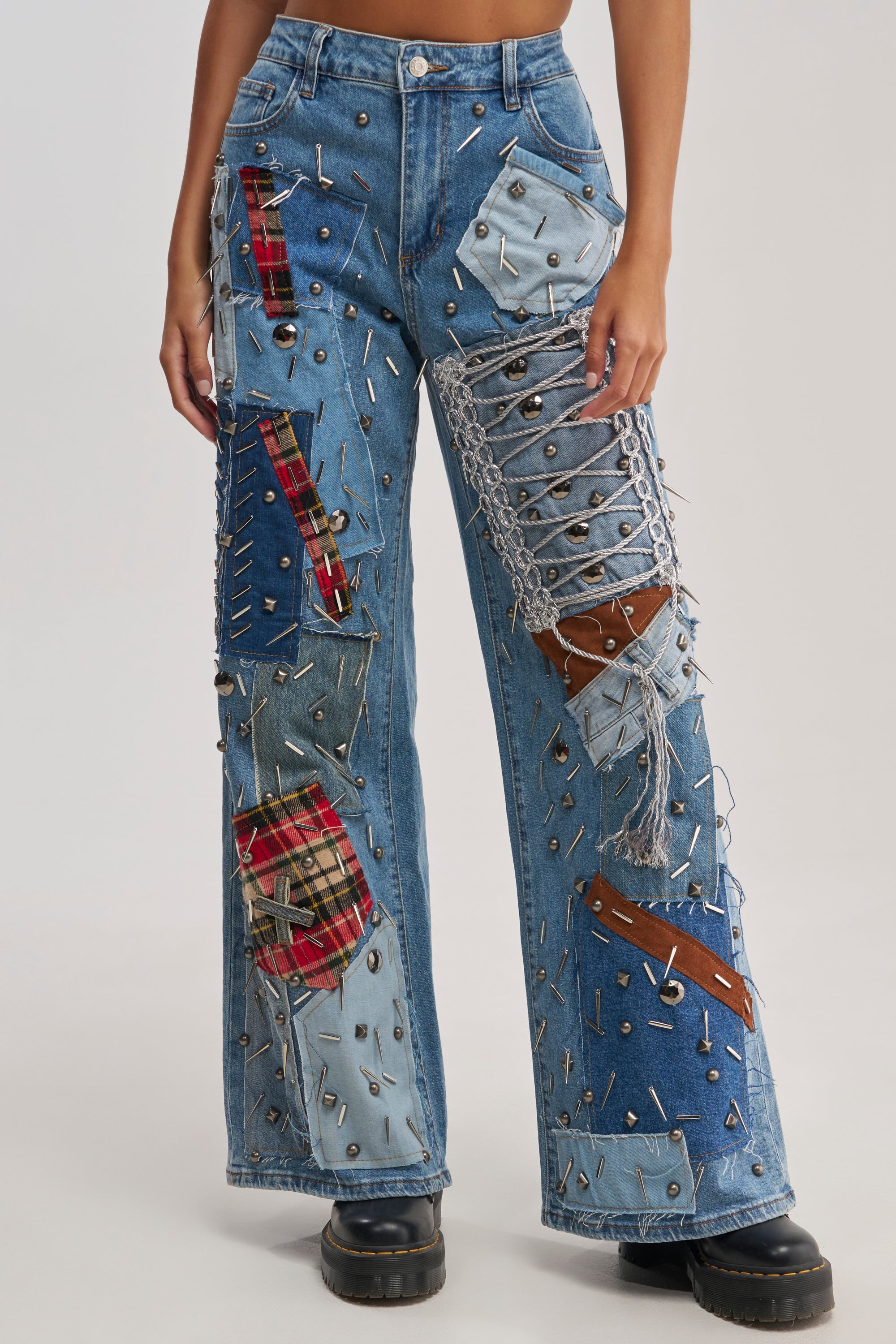 A person wears MIXED EMOTIONS EMBELLISHED DENIM—high-waisted, wide-leg jeans with mixed denim, plaid patches, chains, metal studs, and frayed details—paired with chunky black shoes. The streetwear-inspired photo is cropped at the torso.