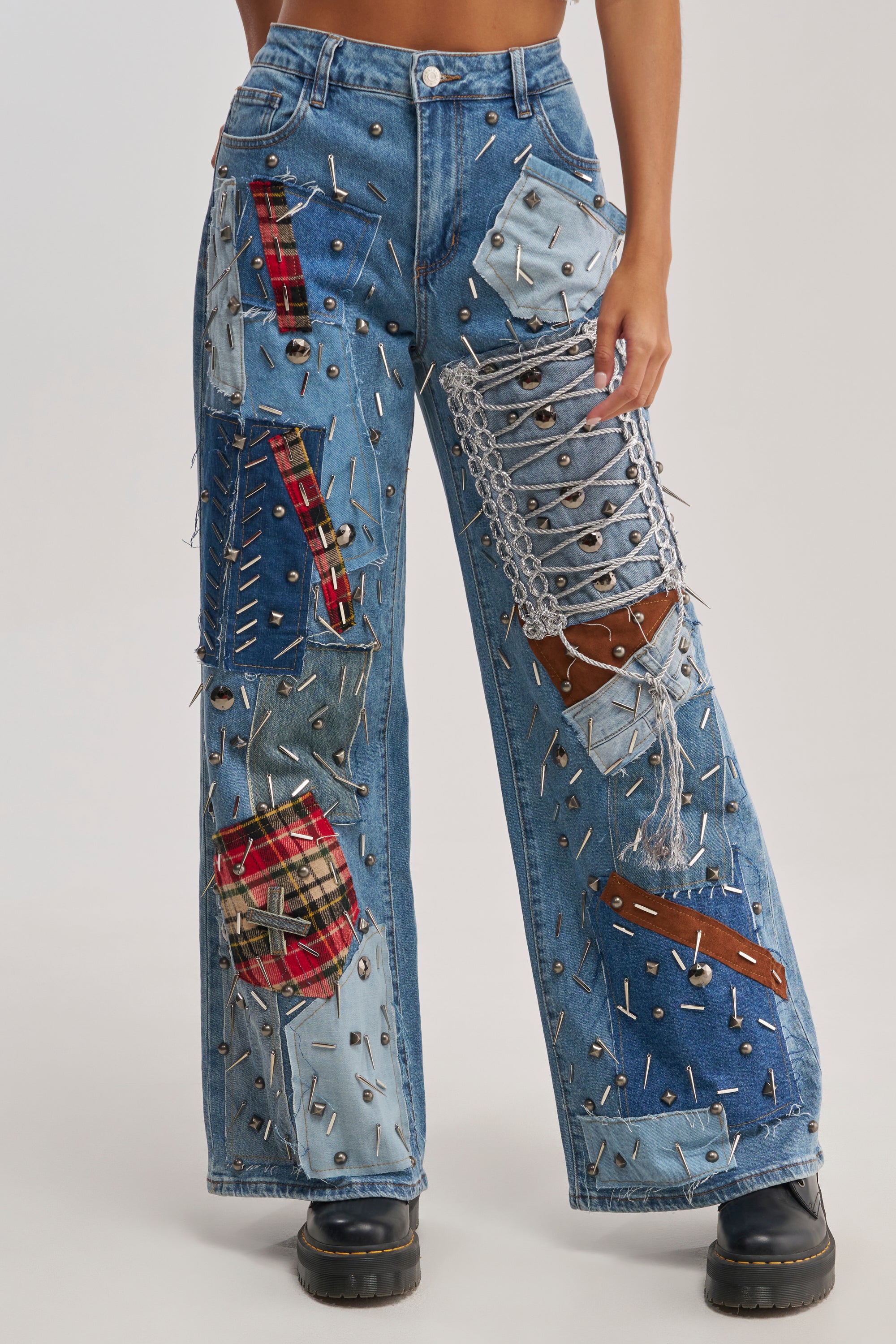 A person wearing MIXED EMOTIONS EMBELLISHED DENIM—wide-leg plaid patch jeans with metal eyelets, chains, safety pins, and a bold punk-inspired design—paired with chunky black boots.