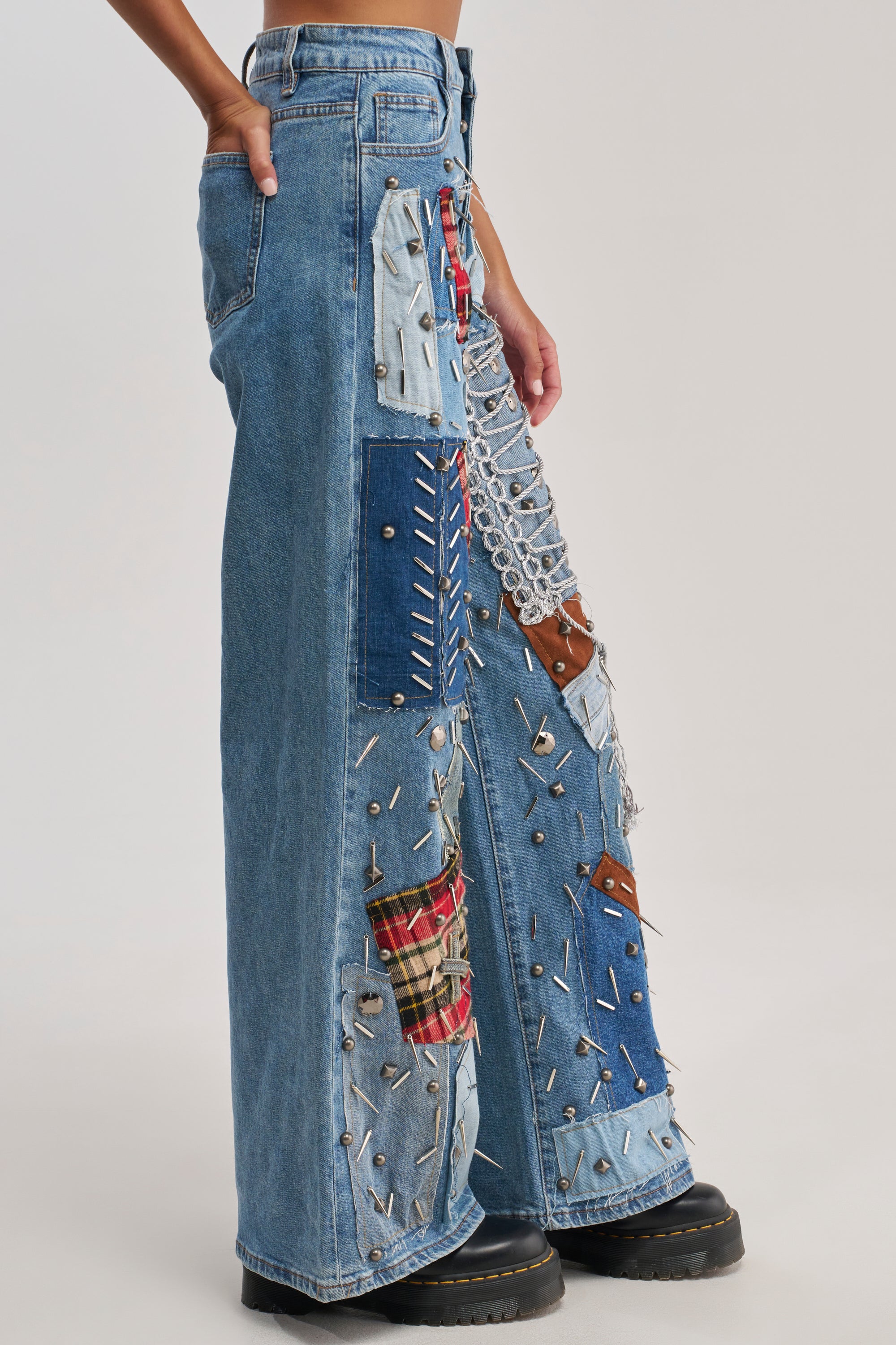 Someone wears the MIXED EMOTIONS EMBELLISHED DENIM—wide-leg jeans with plaid patches, safety pins, and metal chains—paired with black platform boots.
