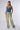 Someone striking a pose against a plain backdrop, dressed strikingly in the REPEAT AFTER ME DENIM PANT, which are wide-legged, light green cargo-style pants with blue accents. They pair this with a standout blue, intricately designed strapless top, accessorized with a chunky necklace and sneakers, showcasing the perfect wide leg silhouette.