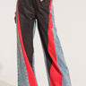 A person is wearing the HARDEST TO LOVE COLORBLOCK DENIM PANT, which boasts a high-rise, wide-legged fit and a striking color-block design with vertical sections of black, red, and denim blue. They complete the look with a bead bracelet and white shoes featuring black tips. The upper body is partially visible.